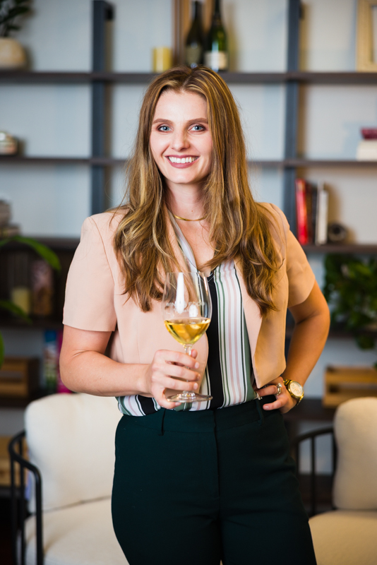 Woman with long hair holding a glass of white wine, smiling, in a modern room with shelves and decor.