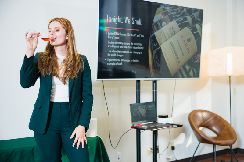 Woman in a suit holding a red pen near her mouth, standing in a room with a TV screen and a chair.