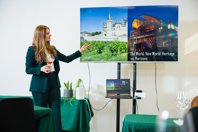 Woman presenting in front of a screen showing two images, one of a castle and one of a modern building, with a laptop on a table nearby.