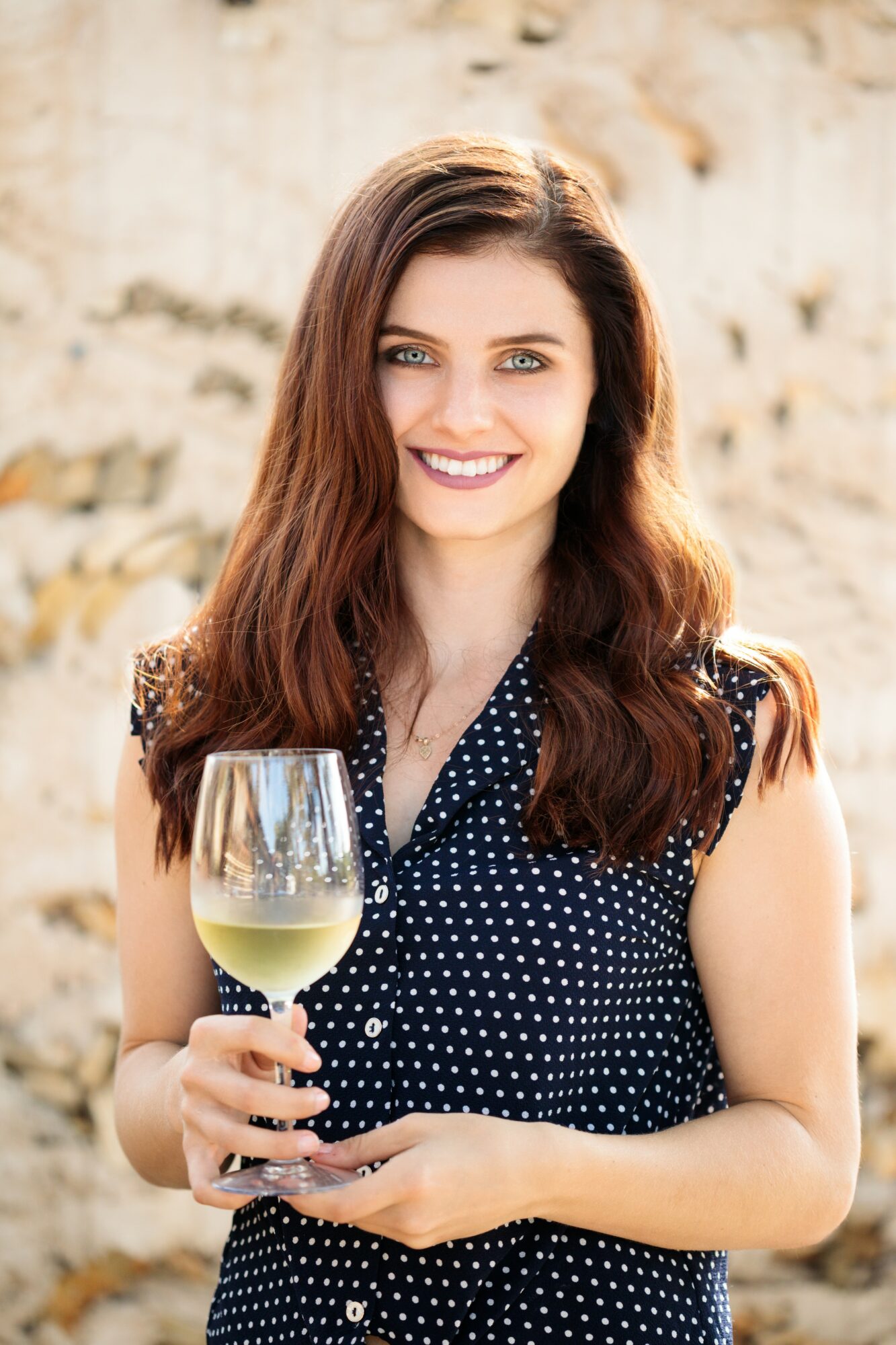 Young woman with long brown hair holding a glass of white wine, smiling outdoors against a stone wall.