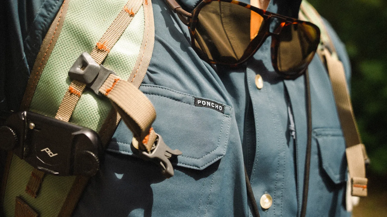 Close-up of a blue backpack with sunglasses hanging on it, outdoors with sunlight and greenery in background.