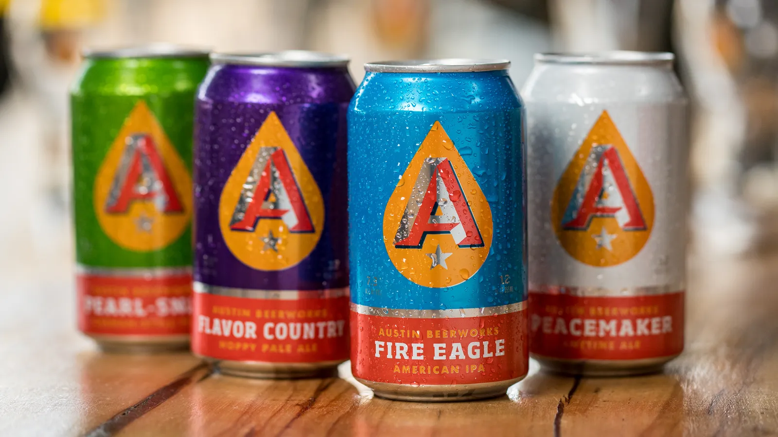 Four cans of beverage with colorful labels on a wooden surface, background blurred, each with a large letter A on the label.