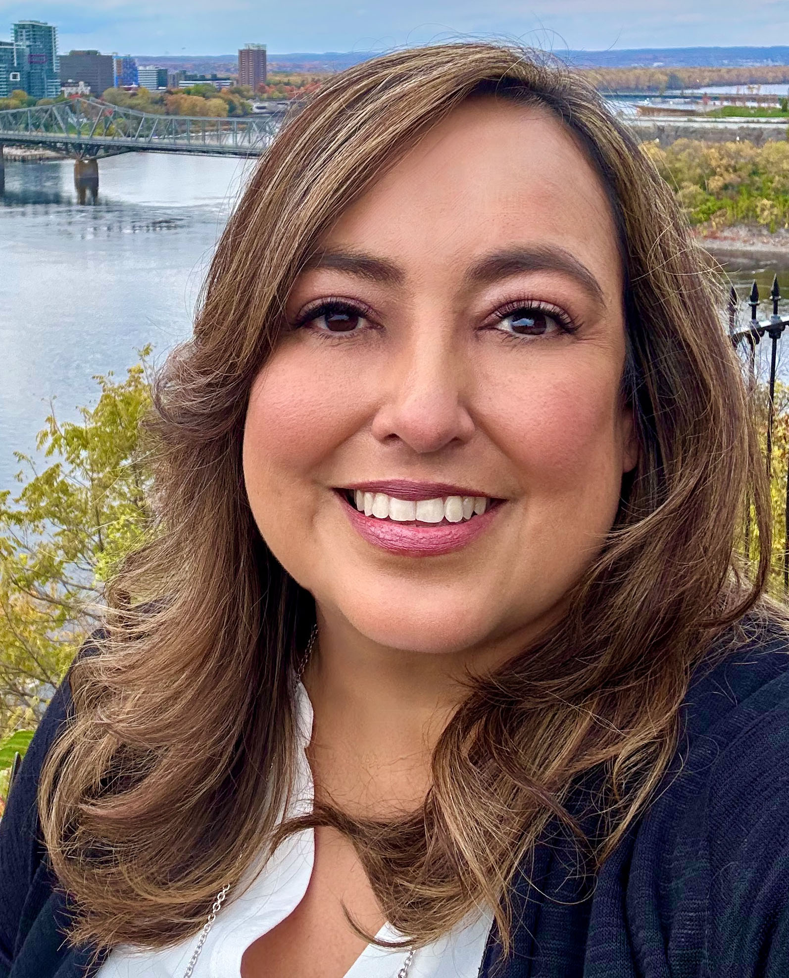 Woman with shoulder-length hair smiling outdoors with a river and cityscape in the background.