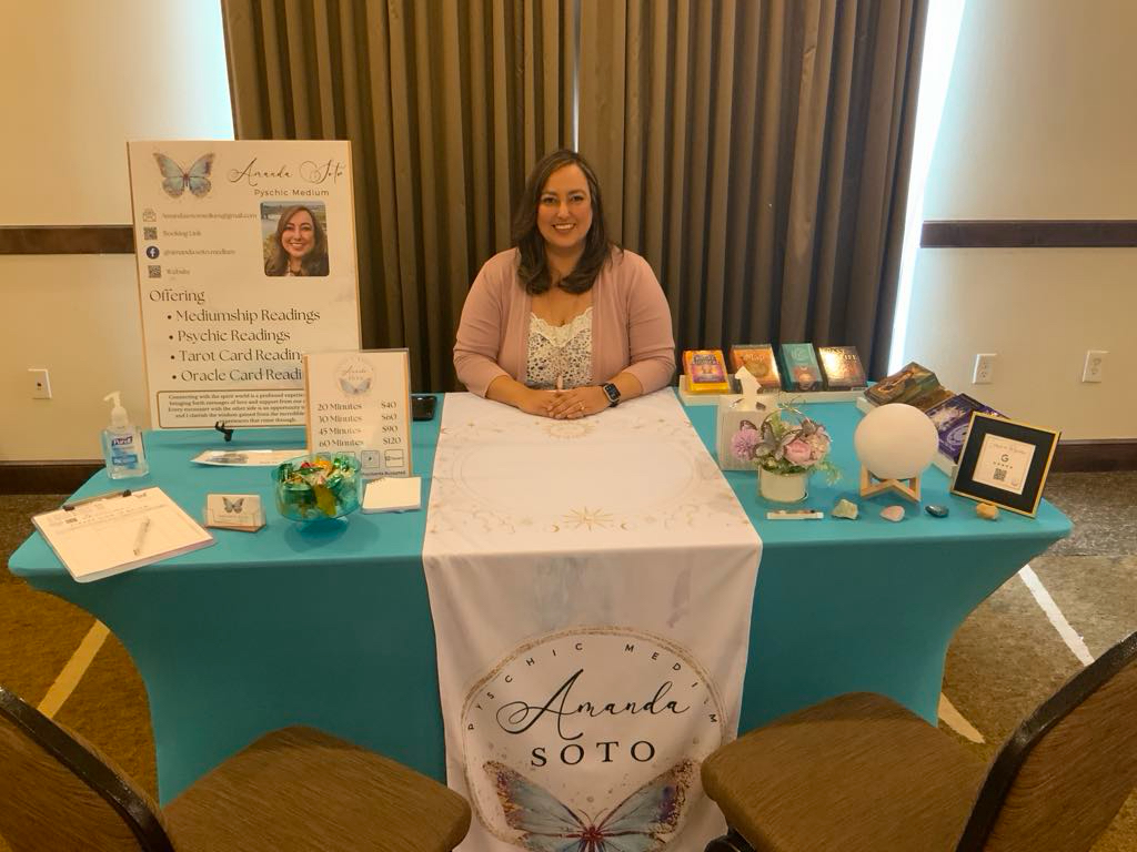 Woman sitting behind a table with promotional materials, a sign, and decorative items, in a room with curtains and a window.