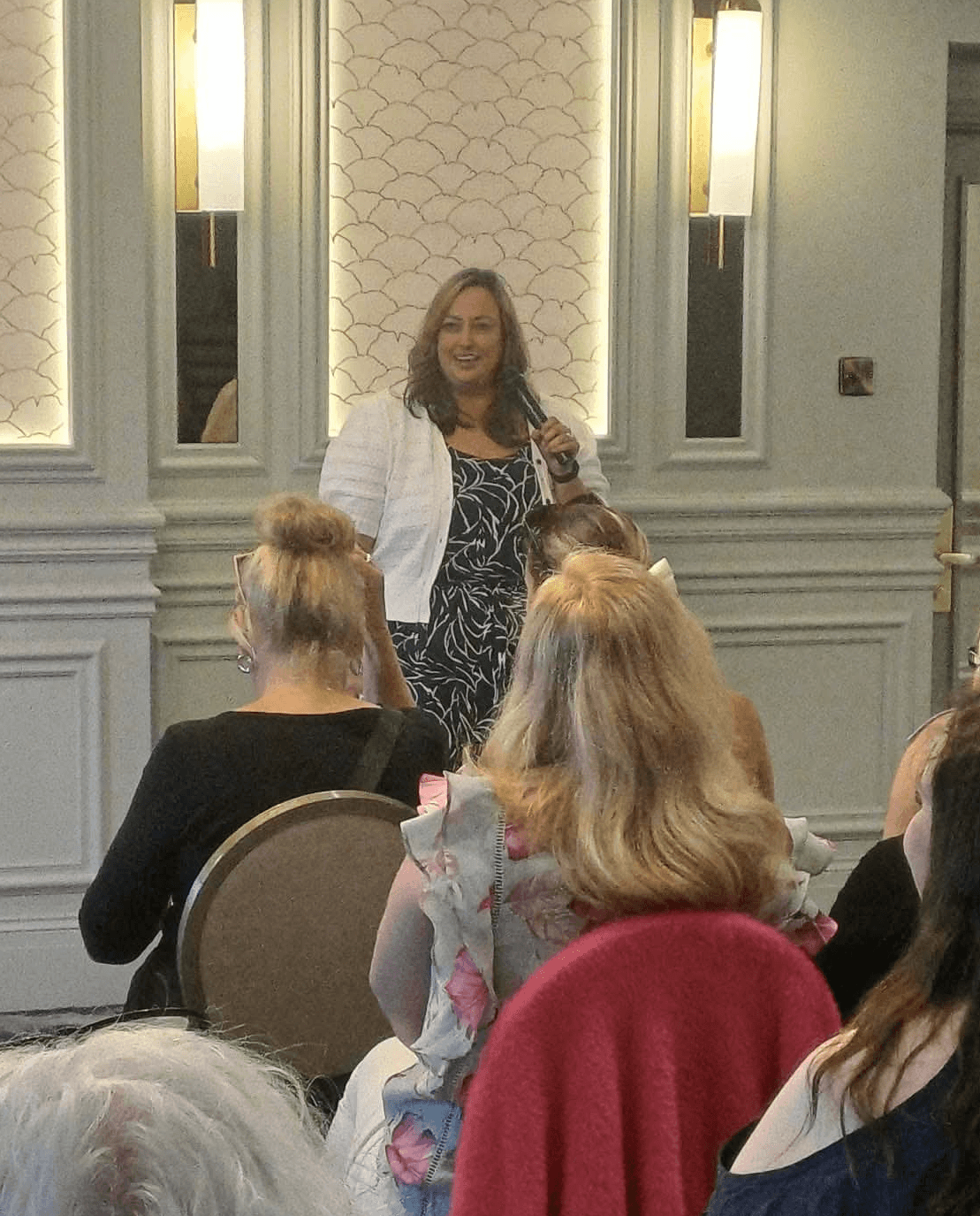 Woman standing and speaking to a seated audience in a room with decorative wall panels and lighting fixtures.