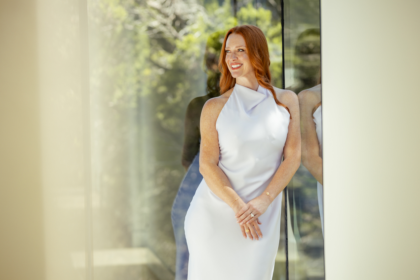 Woman with long red hair in a white sleeveless dress standing indoors near a glass wall, smiling, with greenery outside.