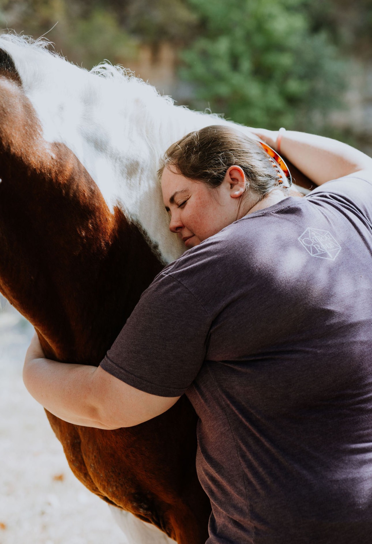 Person hugging a brown and white horse outdoors, with greenery in the background.