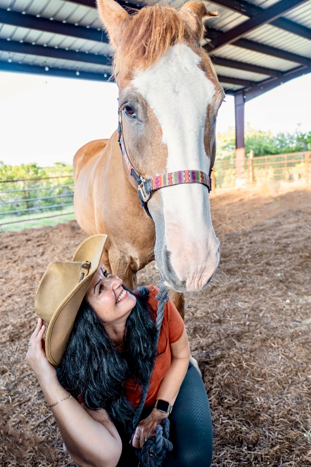 Girl with long dark hair and a hat smiling at a large white and brown horse in a stable.