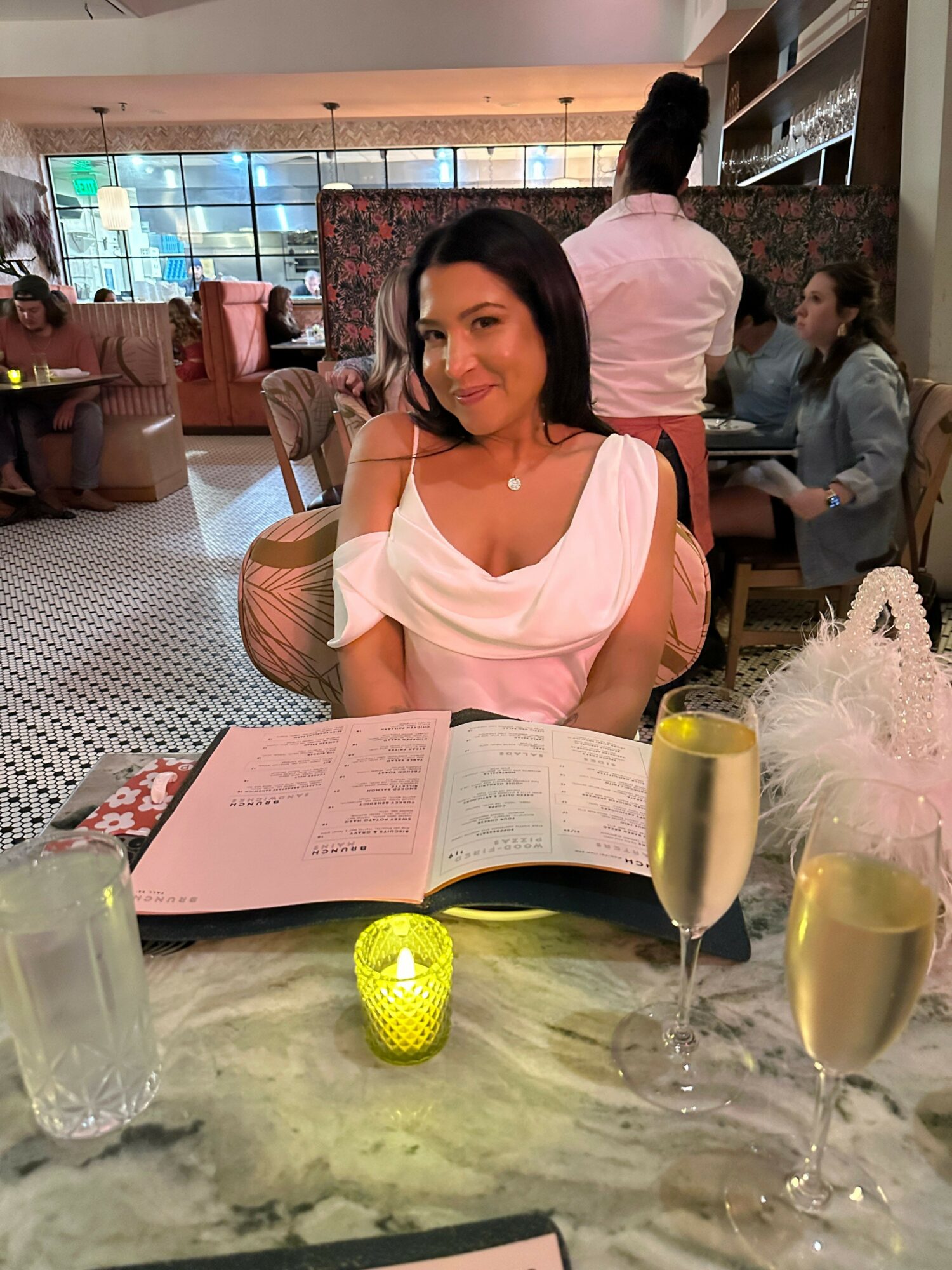 Woman in pink dress reading a menu at a table with drinks, in a lively restaurant setting.