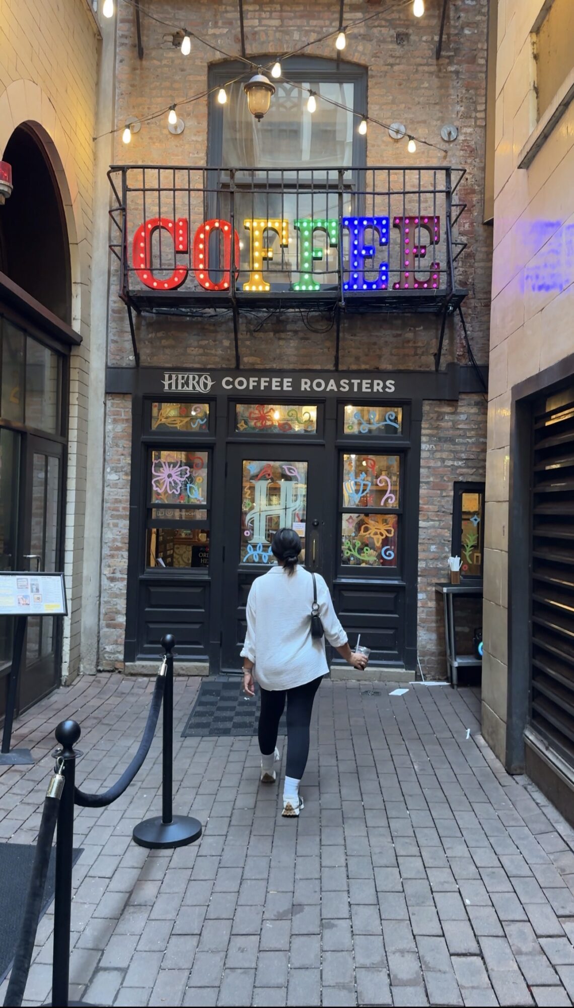Person walking towards a coffee shop entrance with a colorful coffee sign above, brick walls, and string lights.
