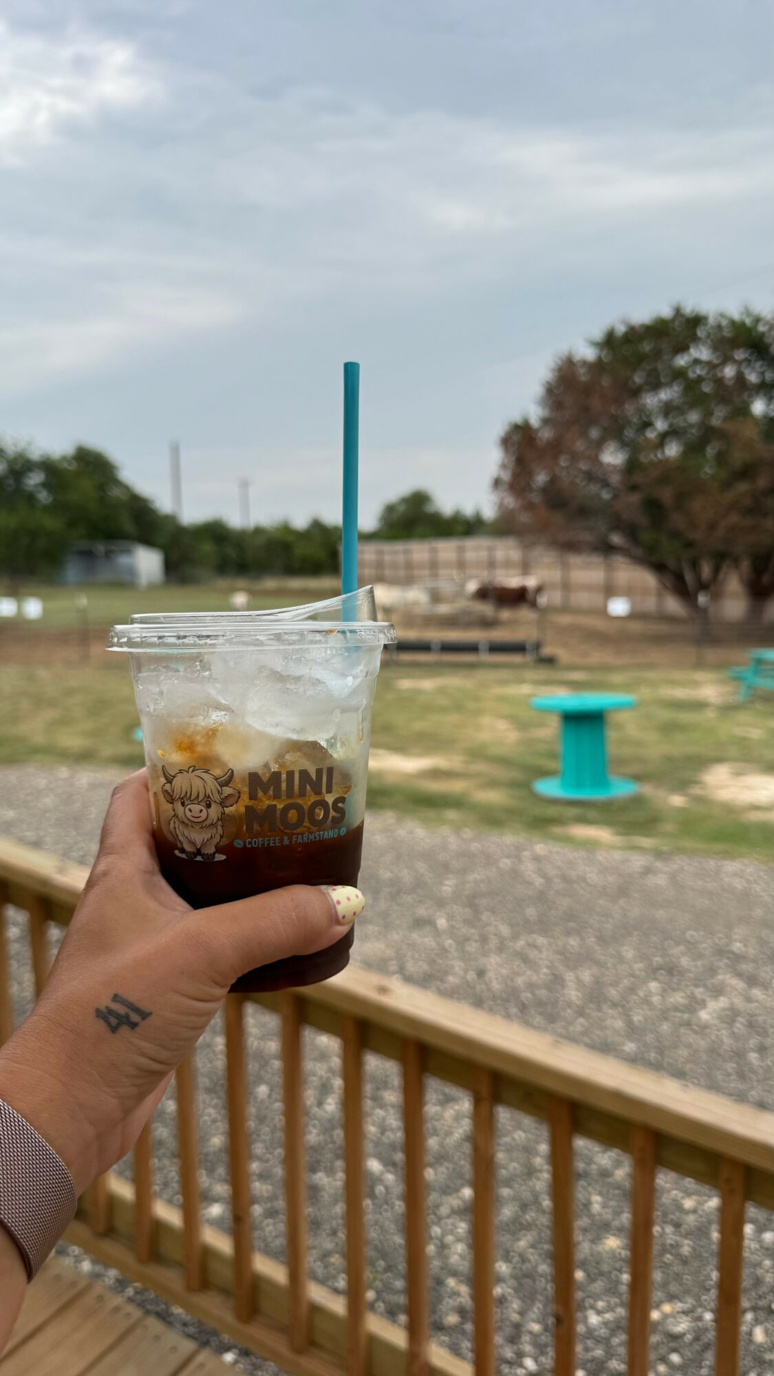 Person holding a plastic cup with a blue straw, outdoors with trees and a blue spool in the background.