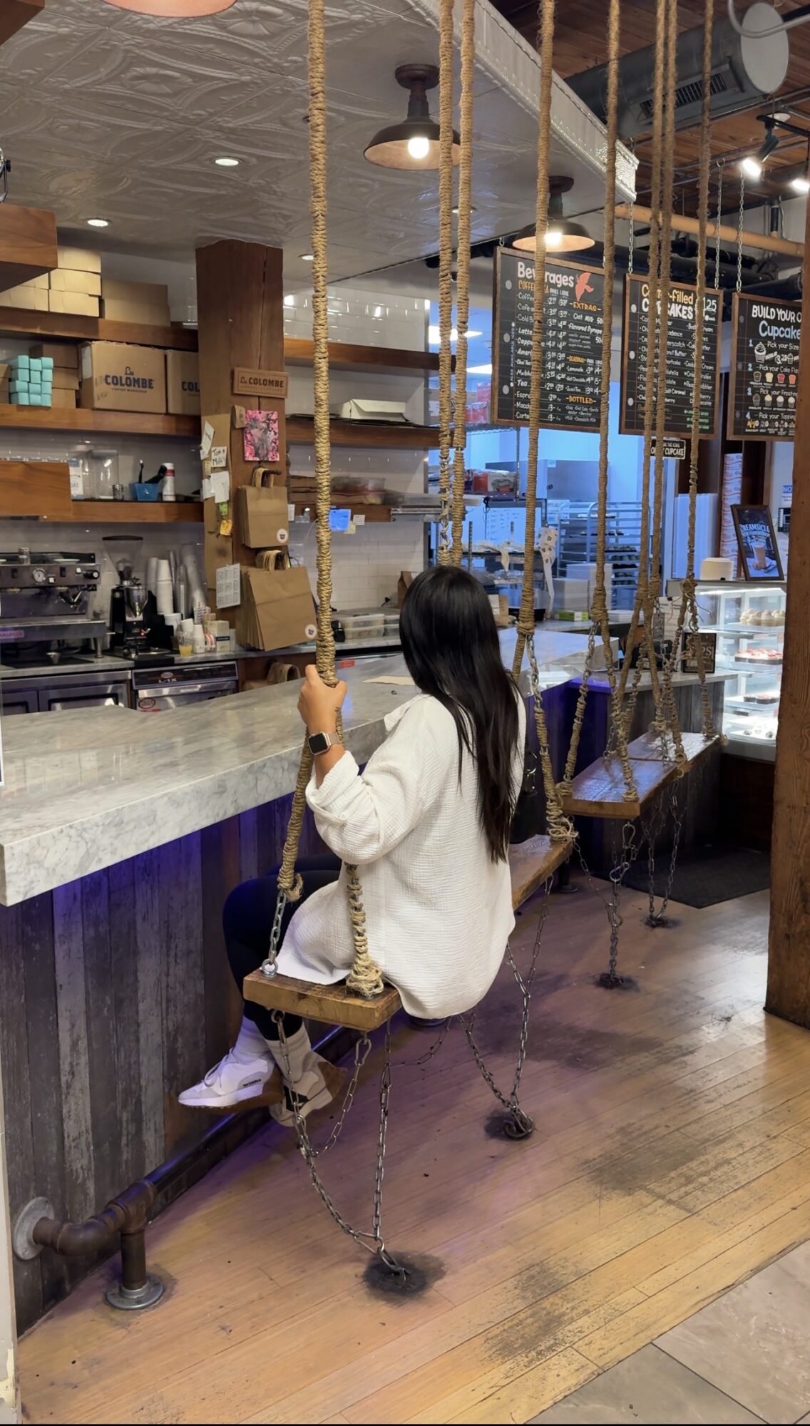 Person sitting on a swing at a cafe counter, facing the menu boards, with a barista area in the background.