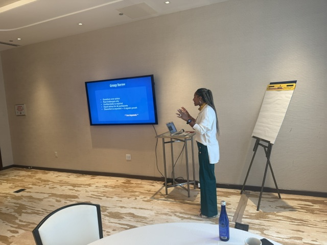 Woman presenting in a conference room with a large screen and flip chart, standing near a table with a laptop and water bottle.