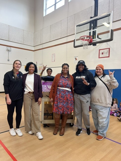 Group of five women standing in a gymnasium near a basketball hoop, smiling and posing for the photo.