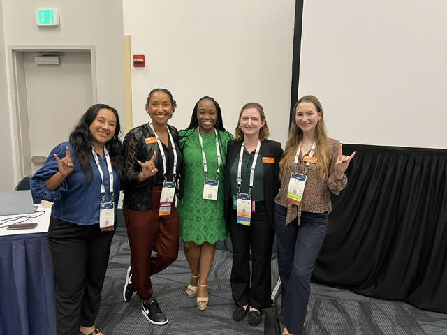 Five women standing together in a conference room, smiling, with name tags and lanyards, posing for a group photo.
