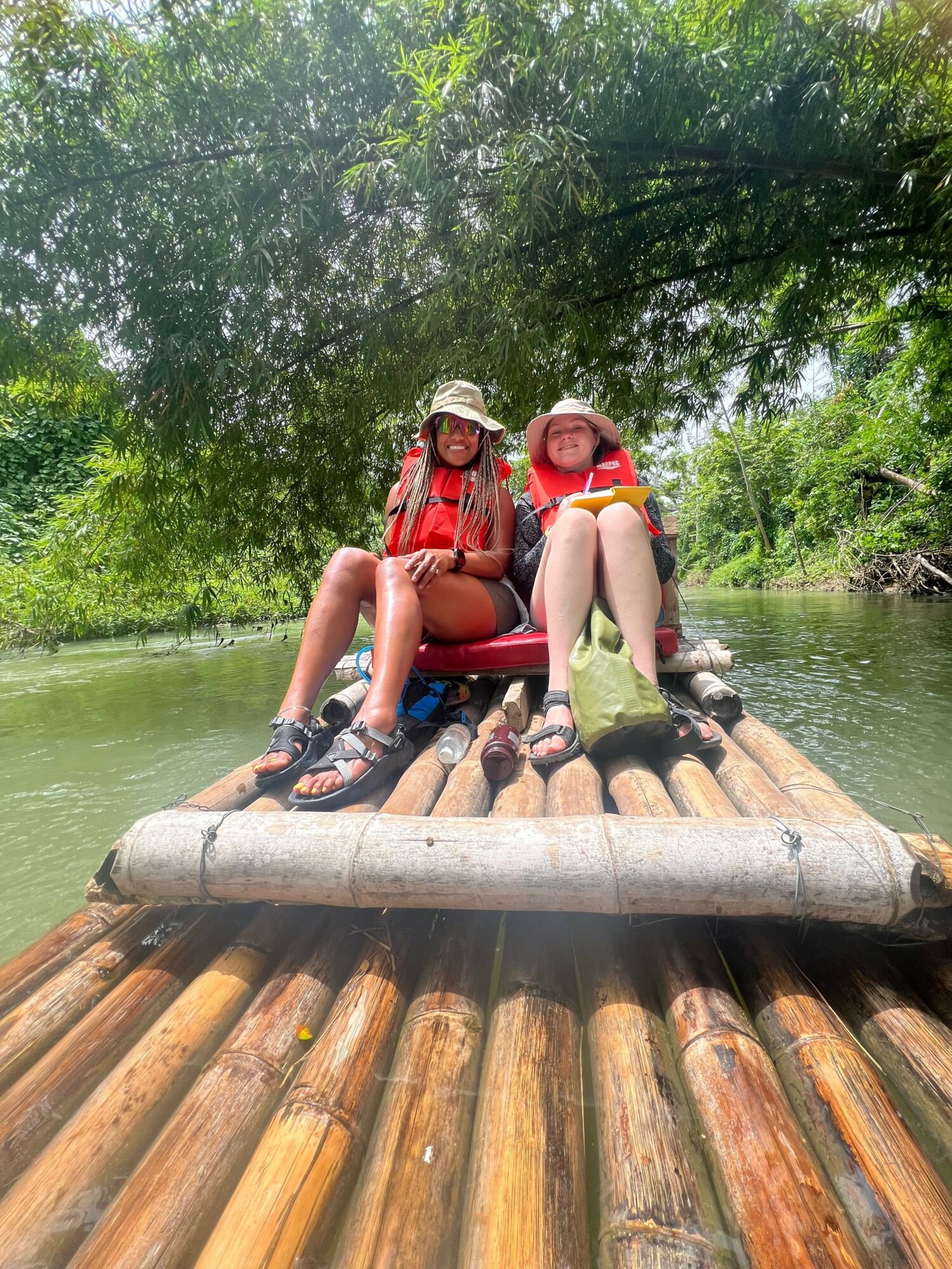 Two people sit on a wooden raft on a river, surrounded by green trees, with one person wearing a red life jacket.