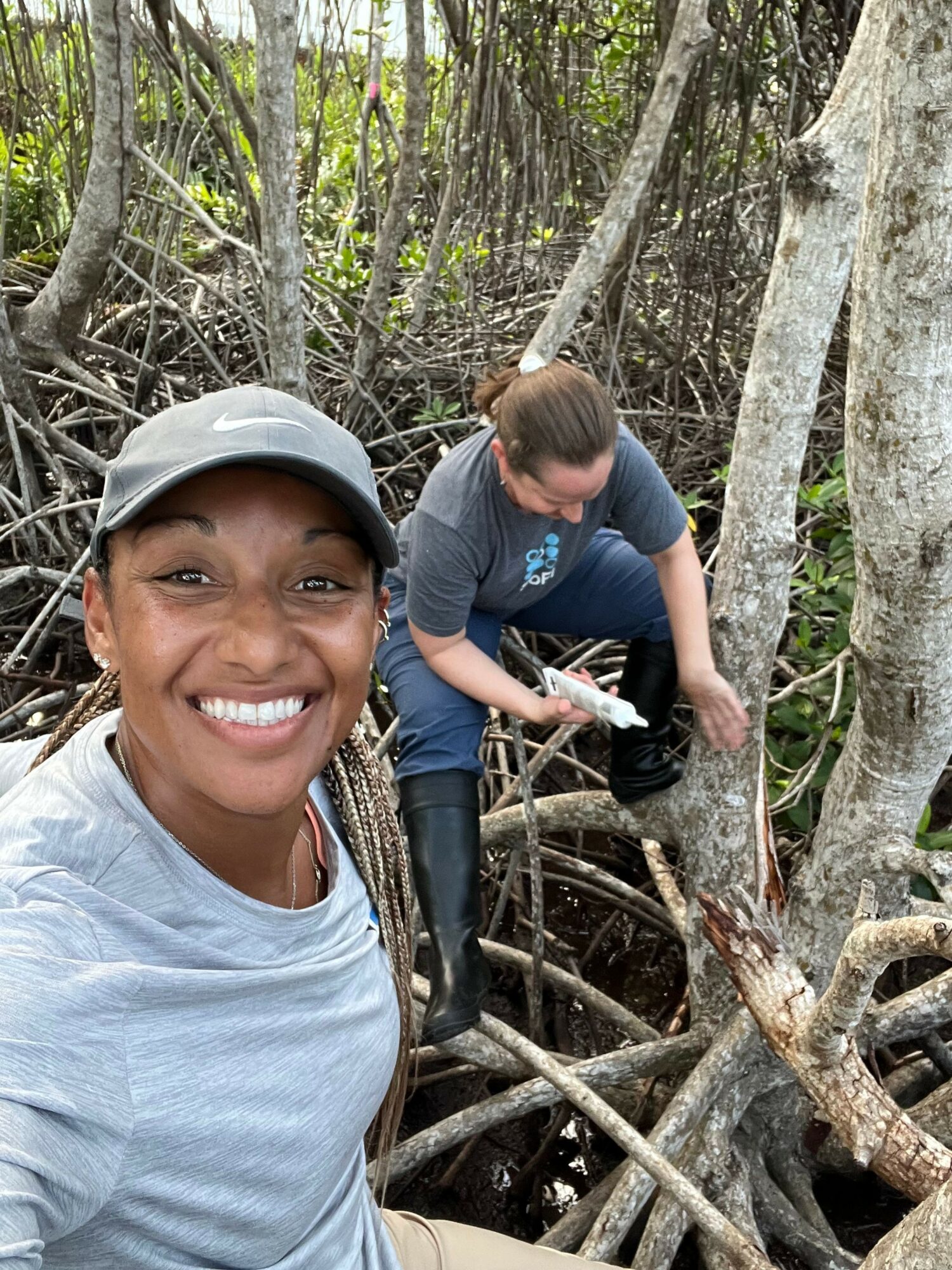 Person smiling in foreground, another person in background among tree roots, holding a notebook, in a wooded area.