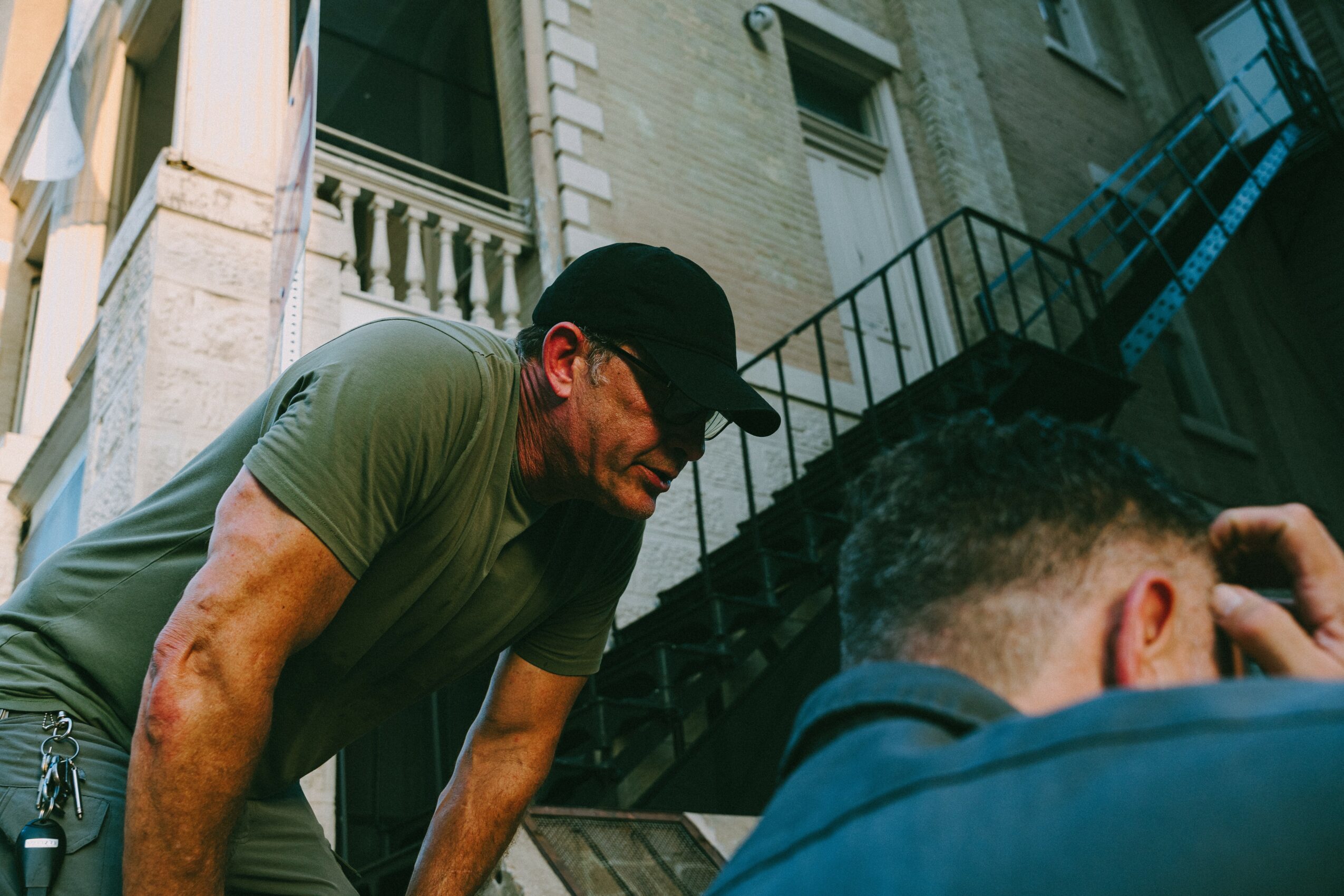 Man wearing a black cap and green shirt leaning forward on a city street, with buildings and stairs in the background.