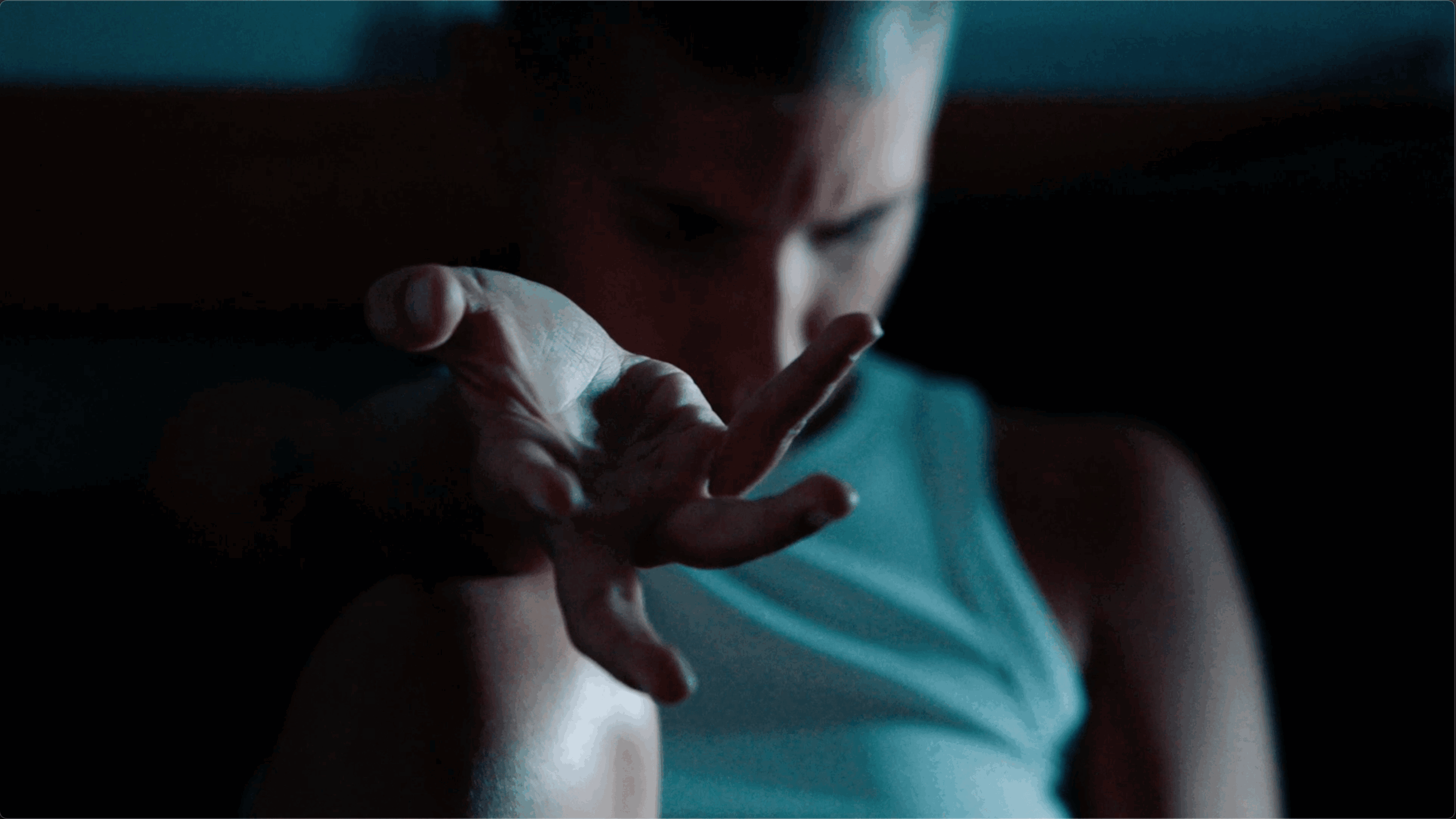 Person reaching out with hand in a dark setting, face partially visible, wearing a sleeveless top.