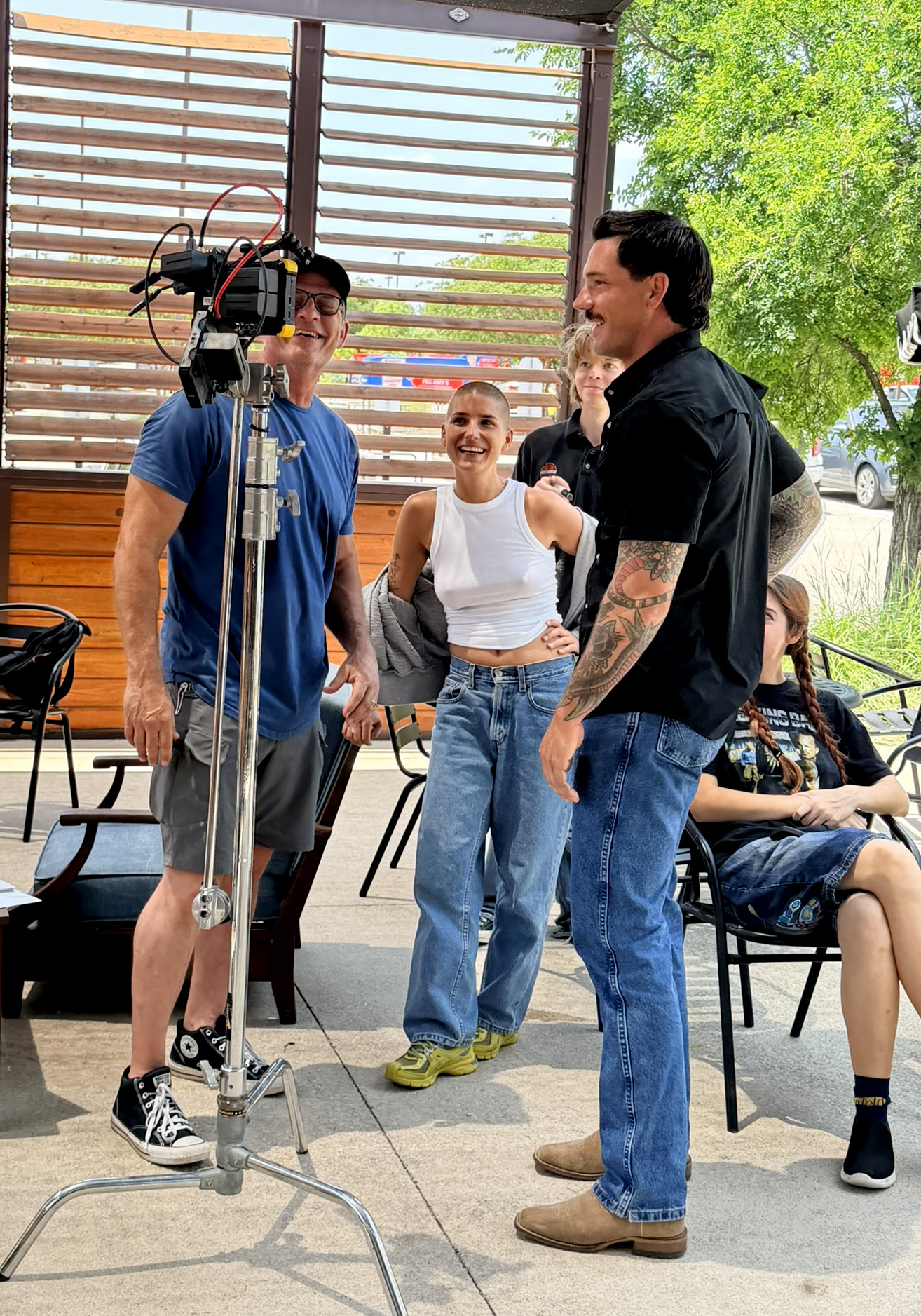 Three people standing and talking outdoors, with a woman sitting nearby, trees and a car in the background.