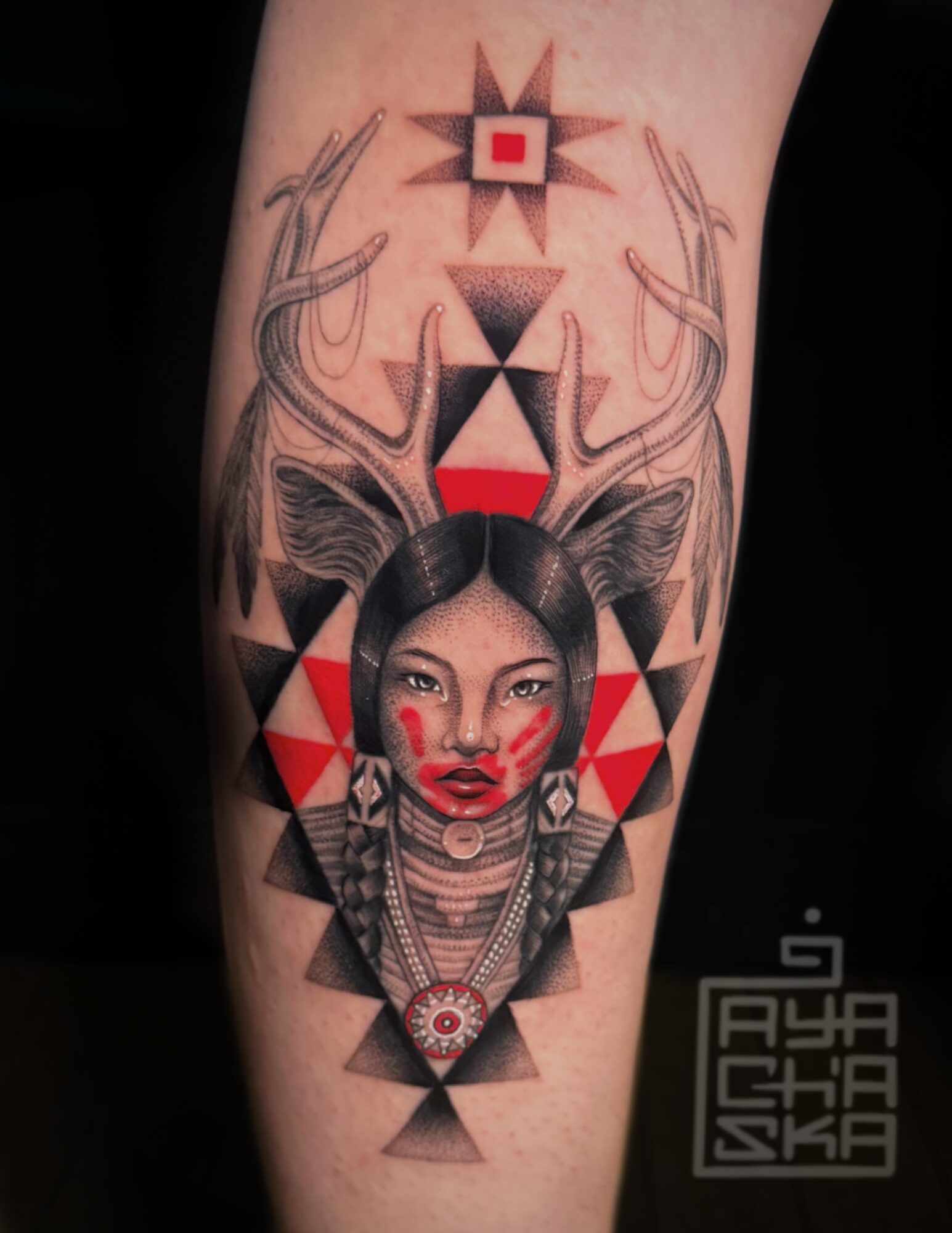Tattoo of a woman with geometric patterns, antlers, and symmetrical designs on arm, with red, black, and gray colors.