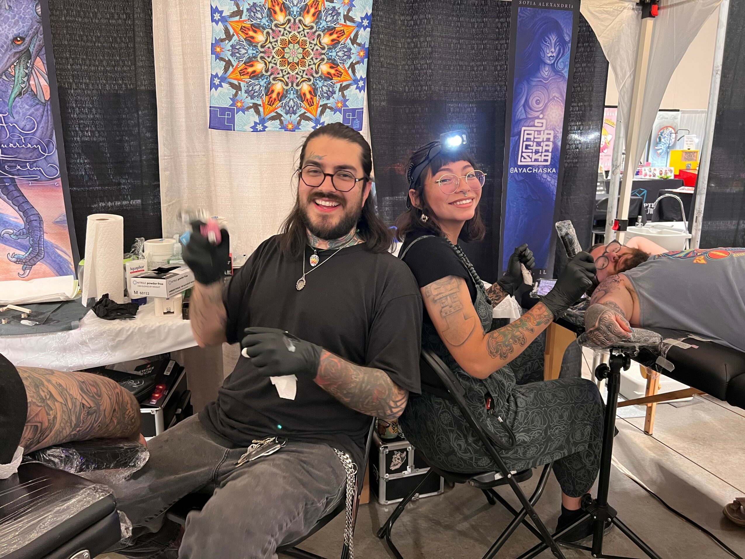 Two tattoo artists smiling and working on clients in a tattoo studio, with colorful artwork in the background.