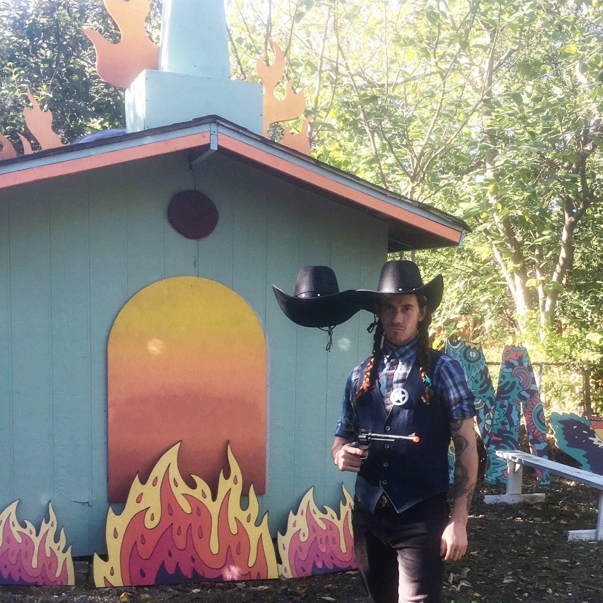 Person wearing a large black hat and vest standing outdoors near a colorful painted structure with flames and sunset scene.
