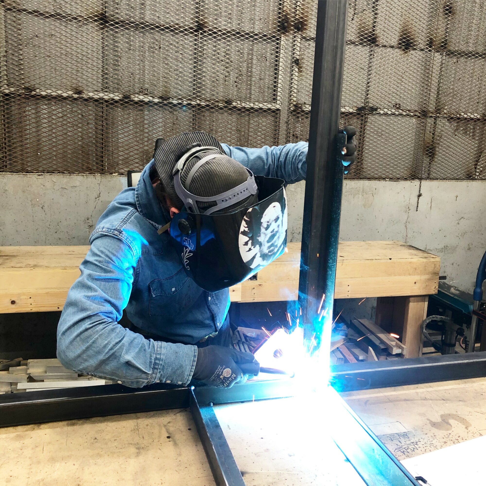 Person welding metal pipe, wearing protective gear, working at a workbench in a workshop.