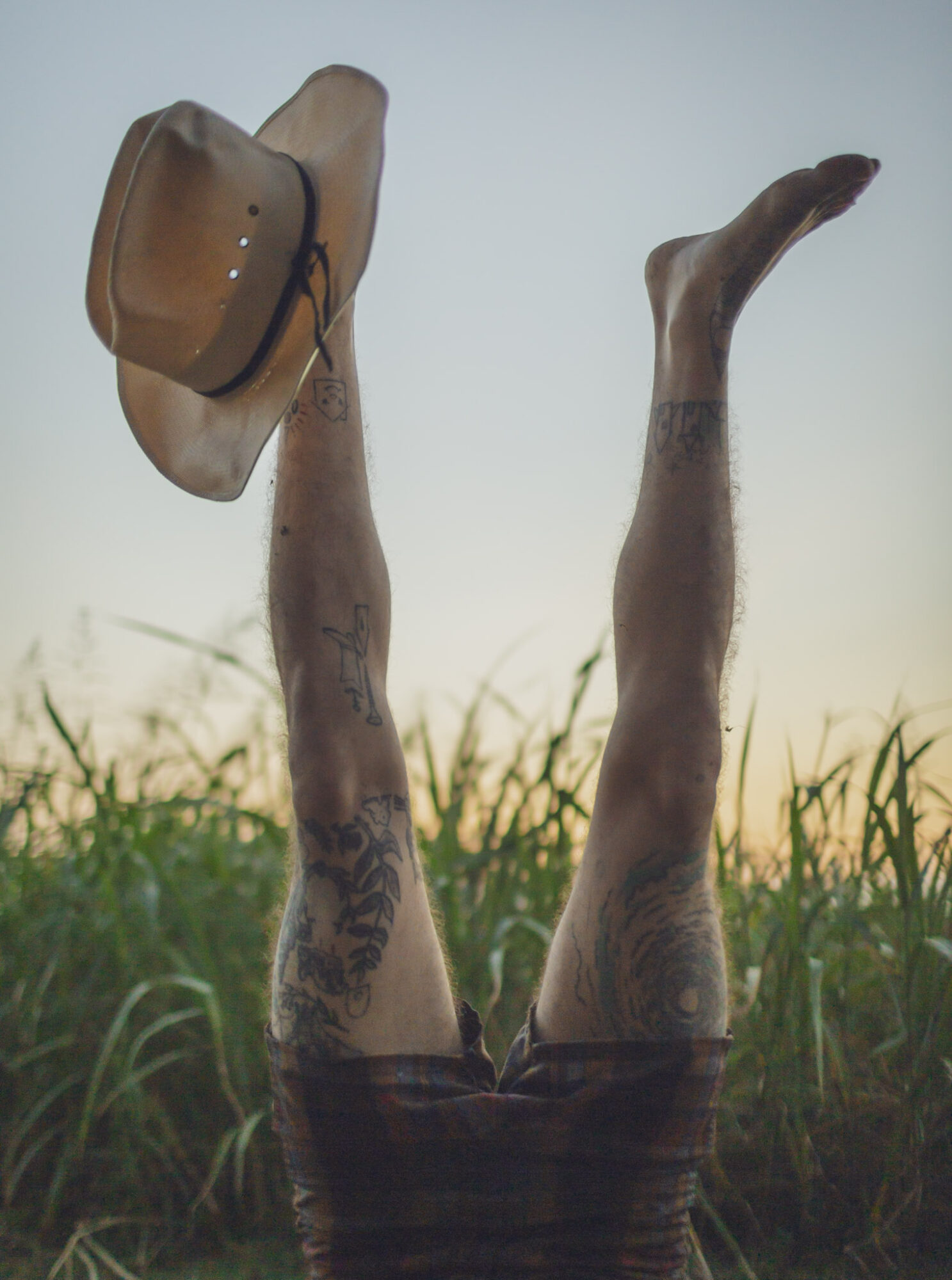 Two tattooed legs raised in the air with a hat hanging from one foot, outdoors with grass and sky background.