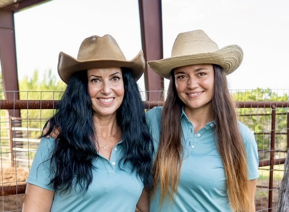 Two women wearing cowboy hats and blue shirts, smiling outdoors with a fence and trees in the background.