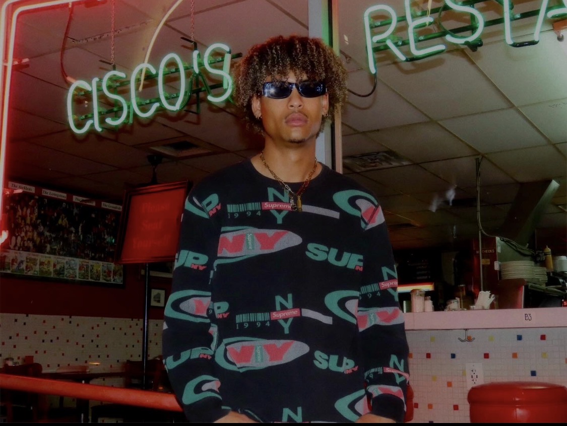 Person with curly hair and sunglasses standing inside a neon-lit restaurant, wearing a patterned sweater.