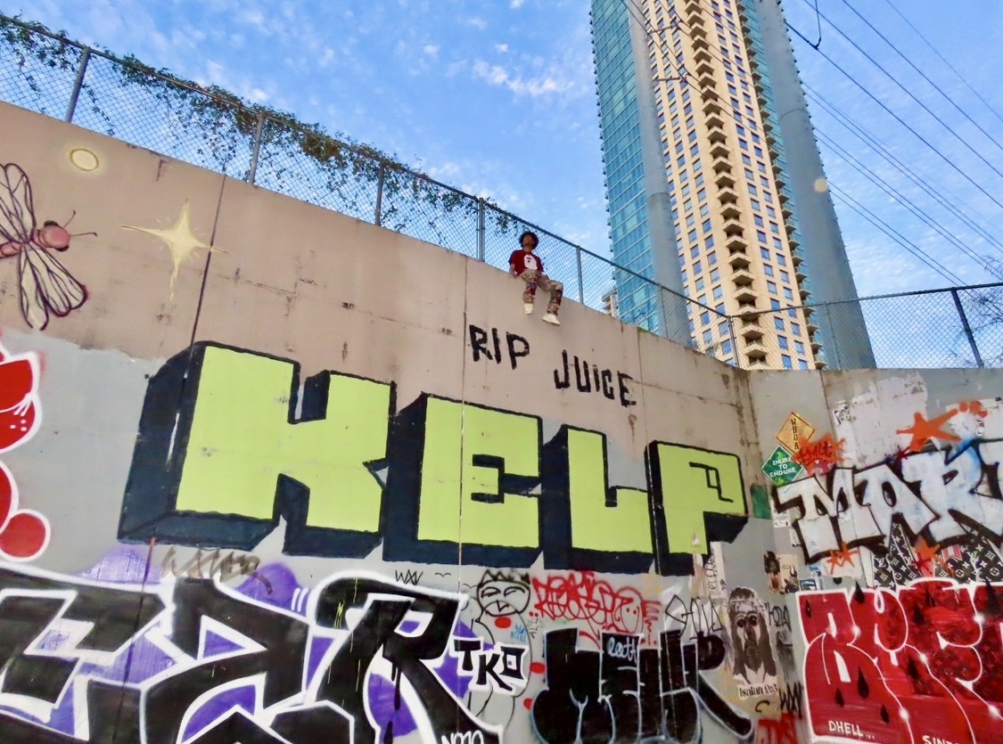 Child climbing a graffiti-covered wall with tall buildings in the background, blue sky, and a fence above the wall.