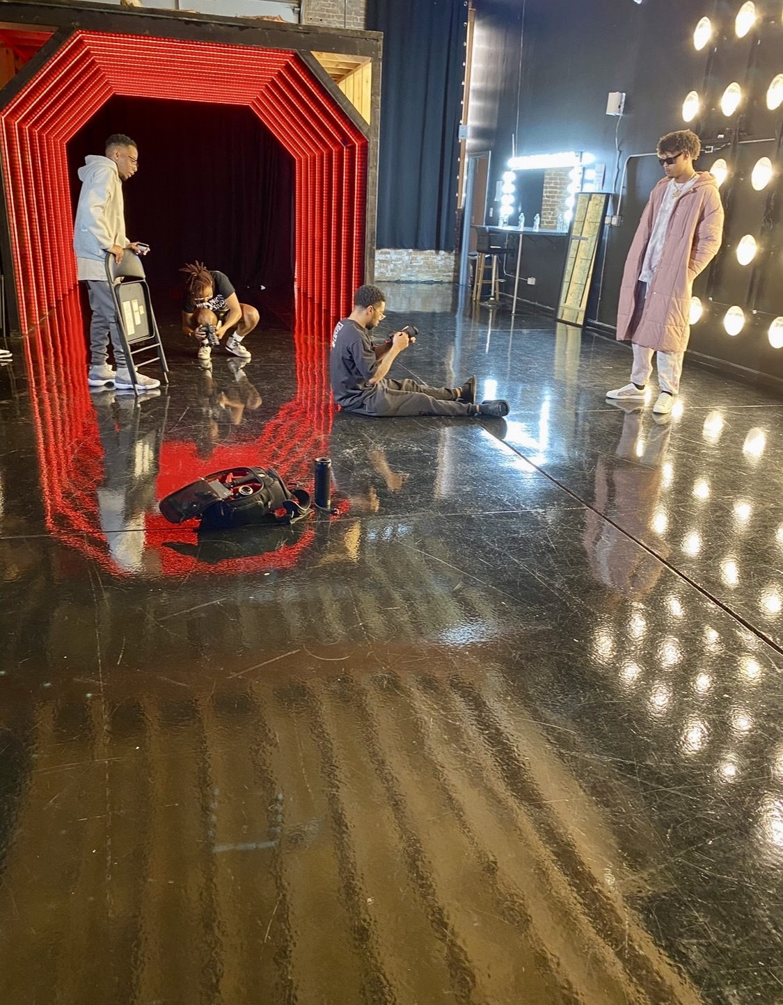 People on a stage with a red arch, some sitting and others standing, with bright lights and a reflective floor.