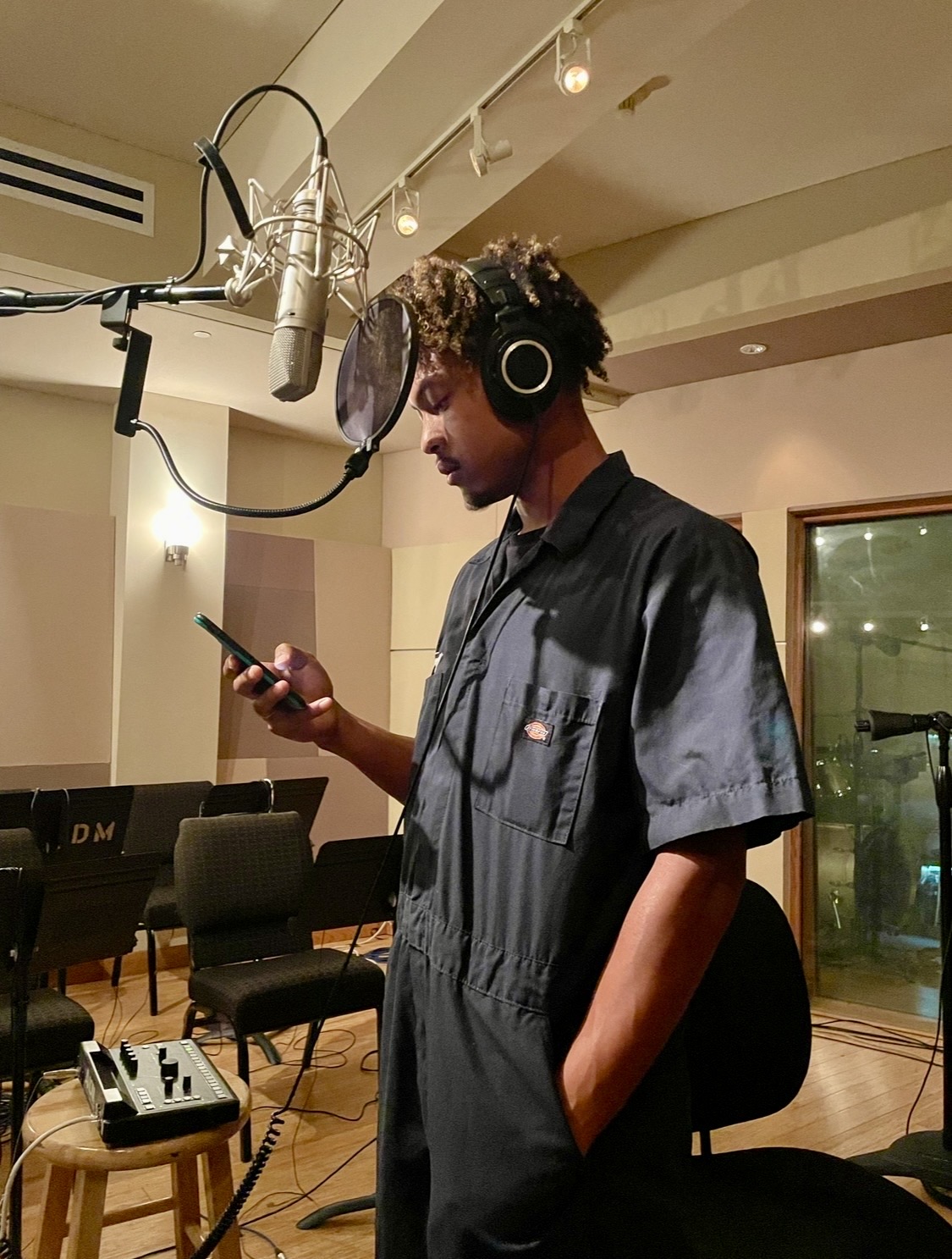Young man with headphones in recording studio, looking at phone, microphone overhead, chairs and window in background.