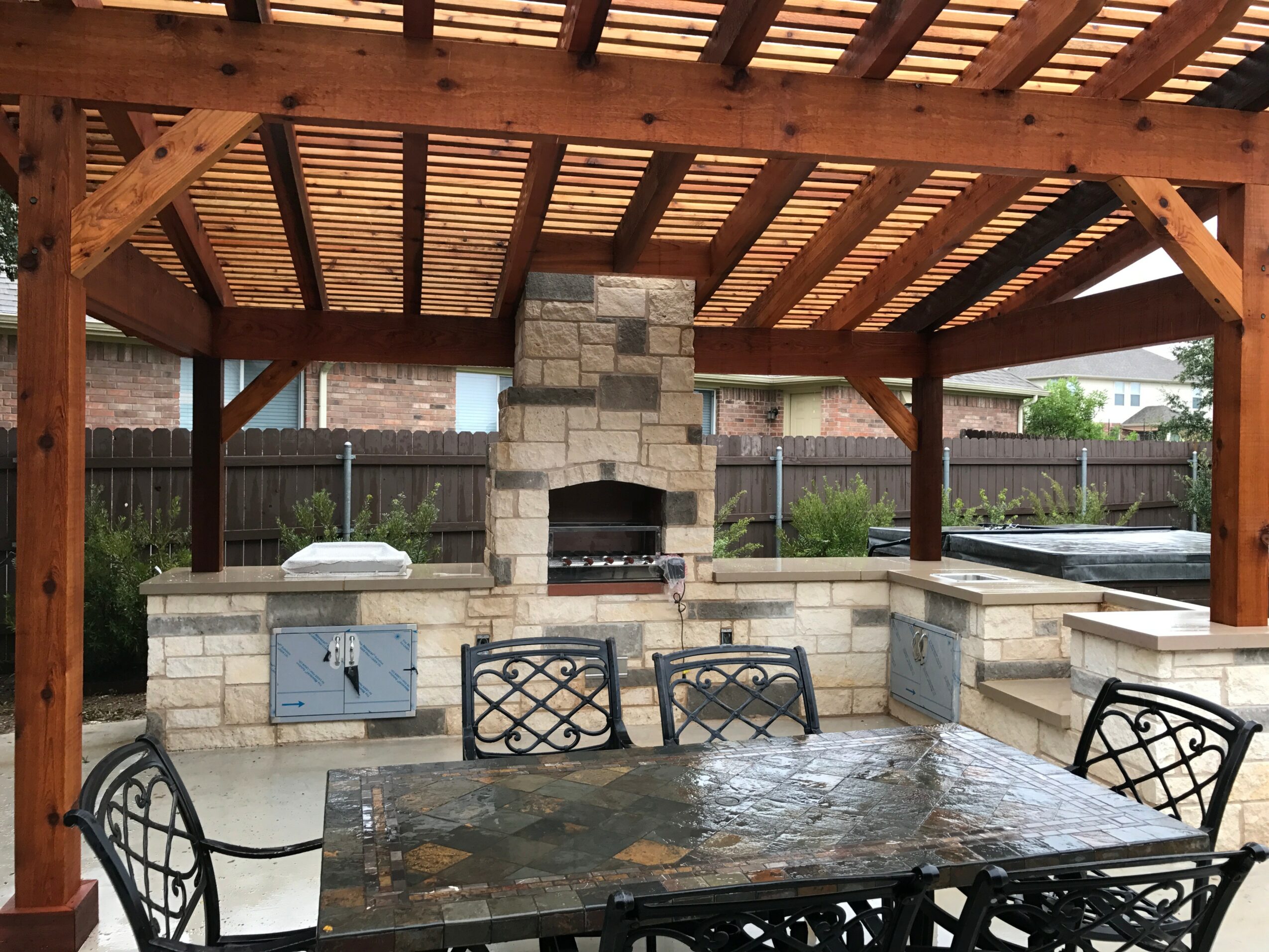 Outdoor dining area with a stone fireplace, wooden roof, and metal chairs around a table.