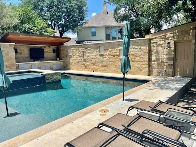Swimming pool with lounge chairs and umbrellas, stone wall, trees, and a house in background.