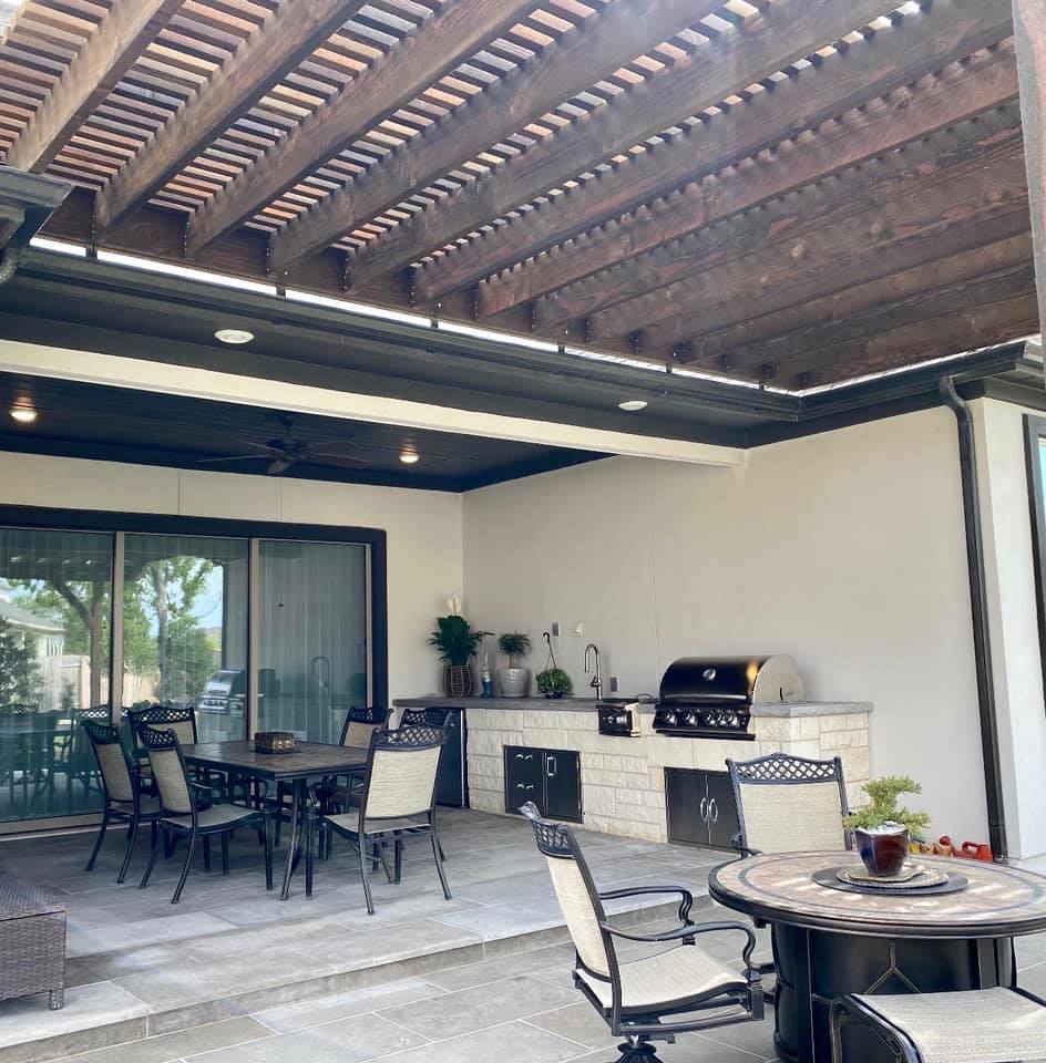 Outdoor dining area with table, chairs, and a grill under a wooden pergola, with a sliding glass door and potted plants.