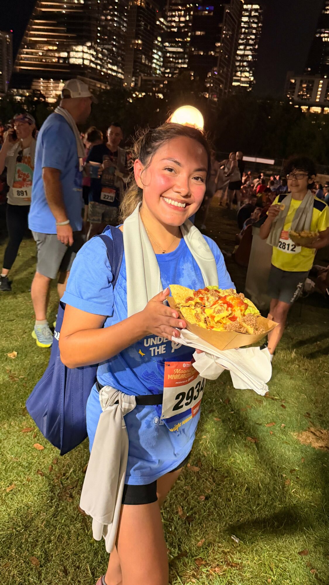 Smiling woman holding a pizza at an outdoor event during nighttime, with city buildings in the background.