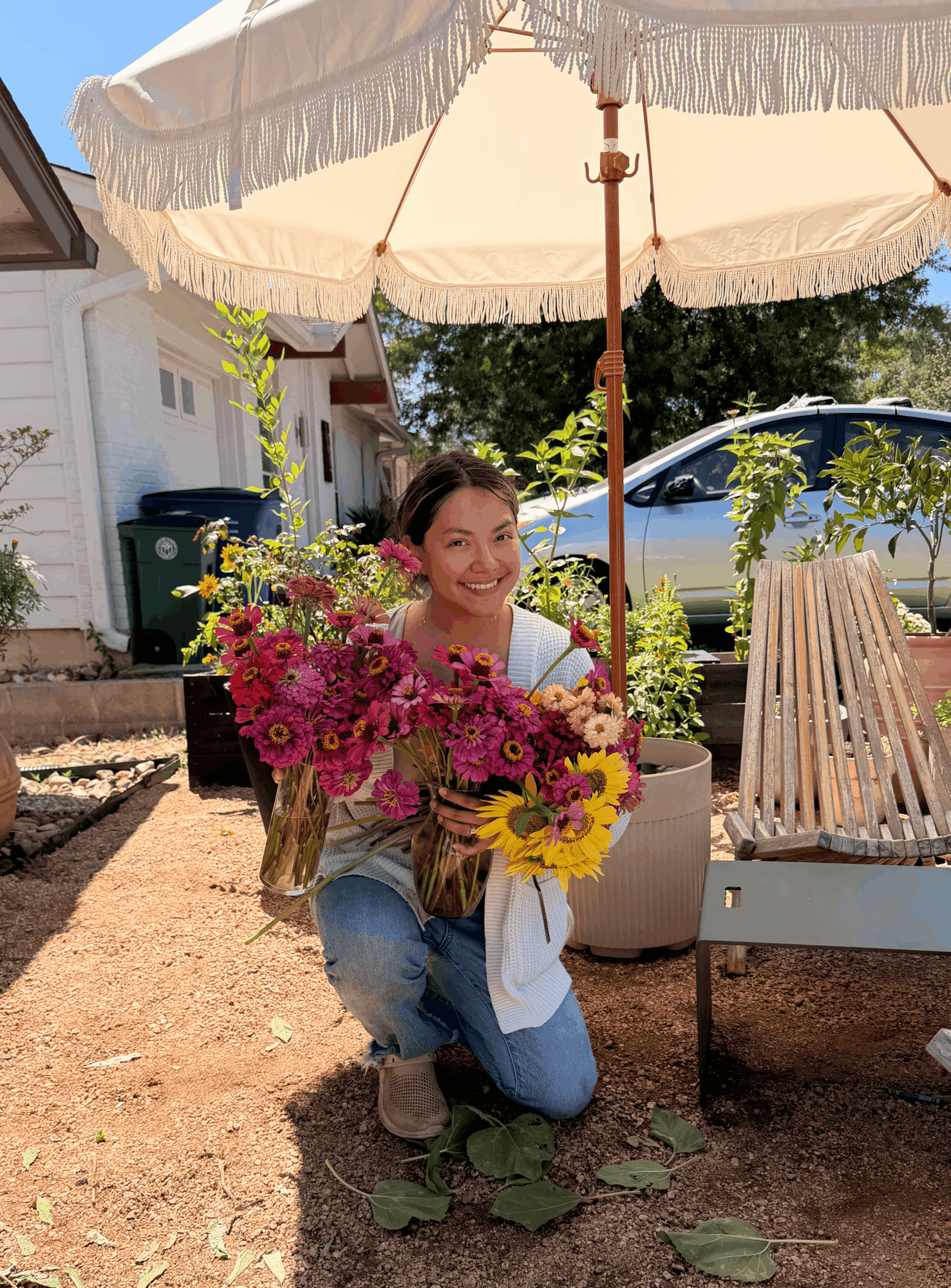 Young woman kneeling outdoors under a large white umbrella, holding colorful flowers, smiling, with a house and car in background.