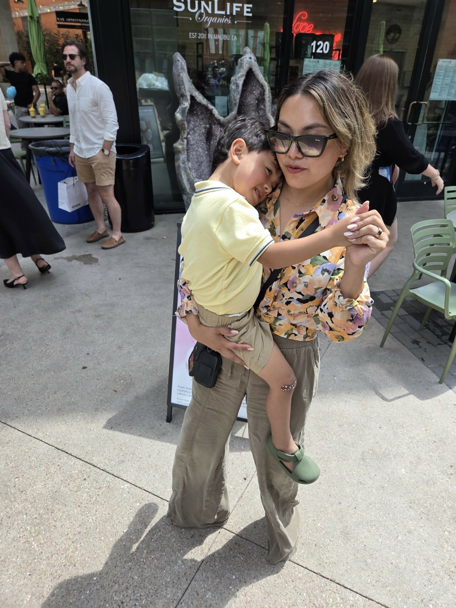 Woman holding a child in a yellow shirt and glasses, standing outdoors with people and chairs around.