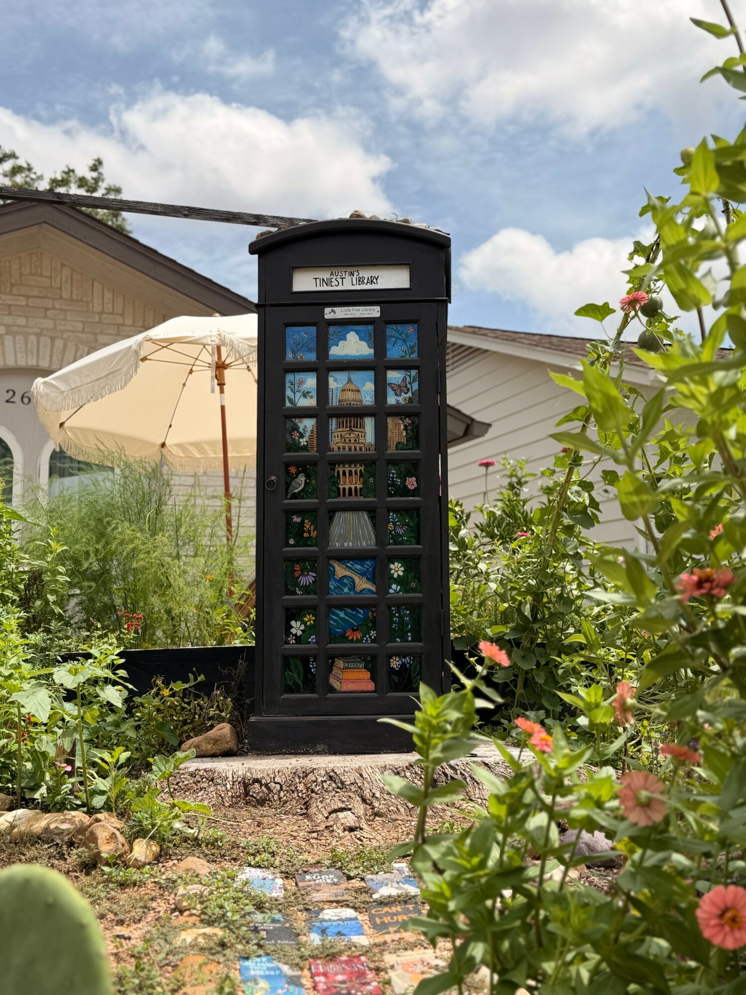 Black telephone booth with glass panels in a garden with plants and flowers, under a partly cloudy sky.