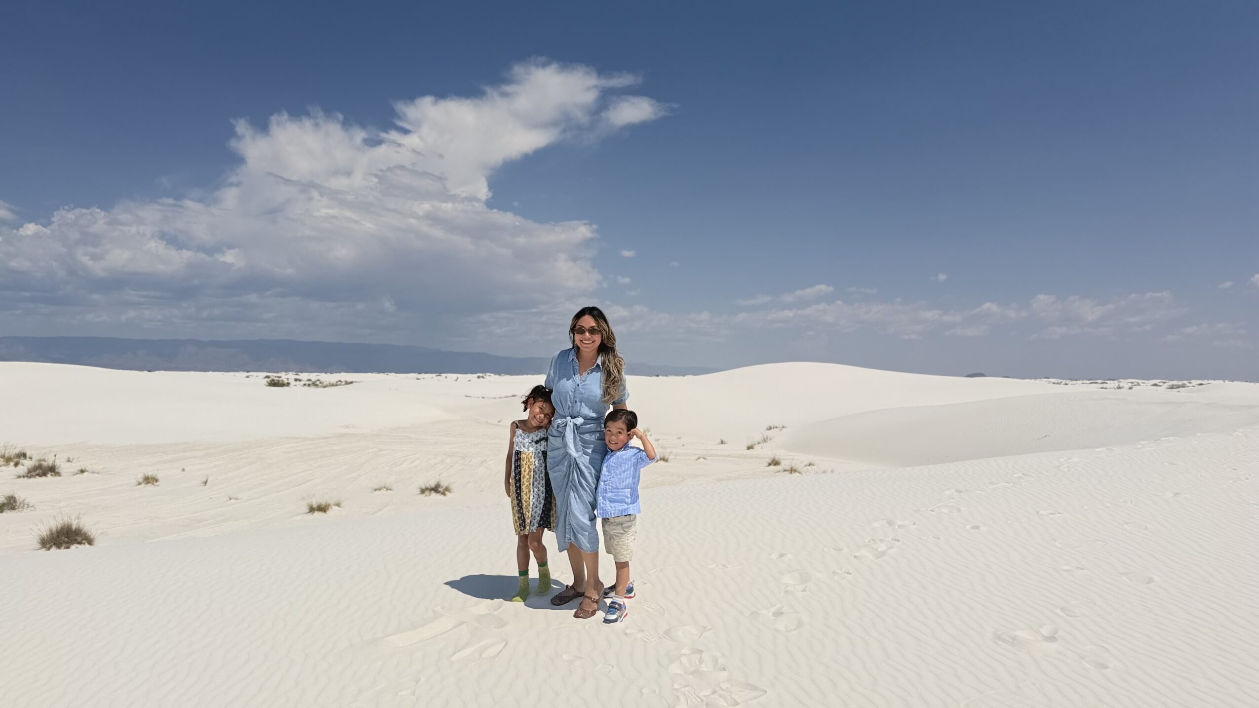 Three people standing on white sand dunes under a blue sky with clouds.