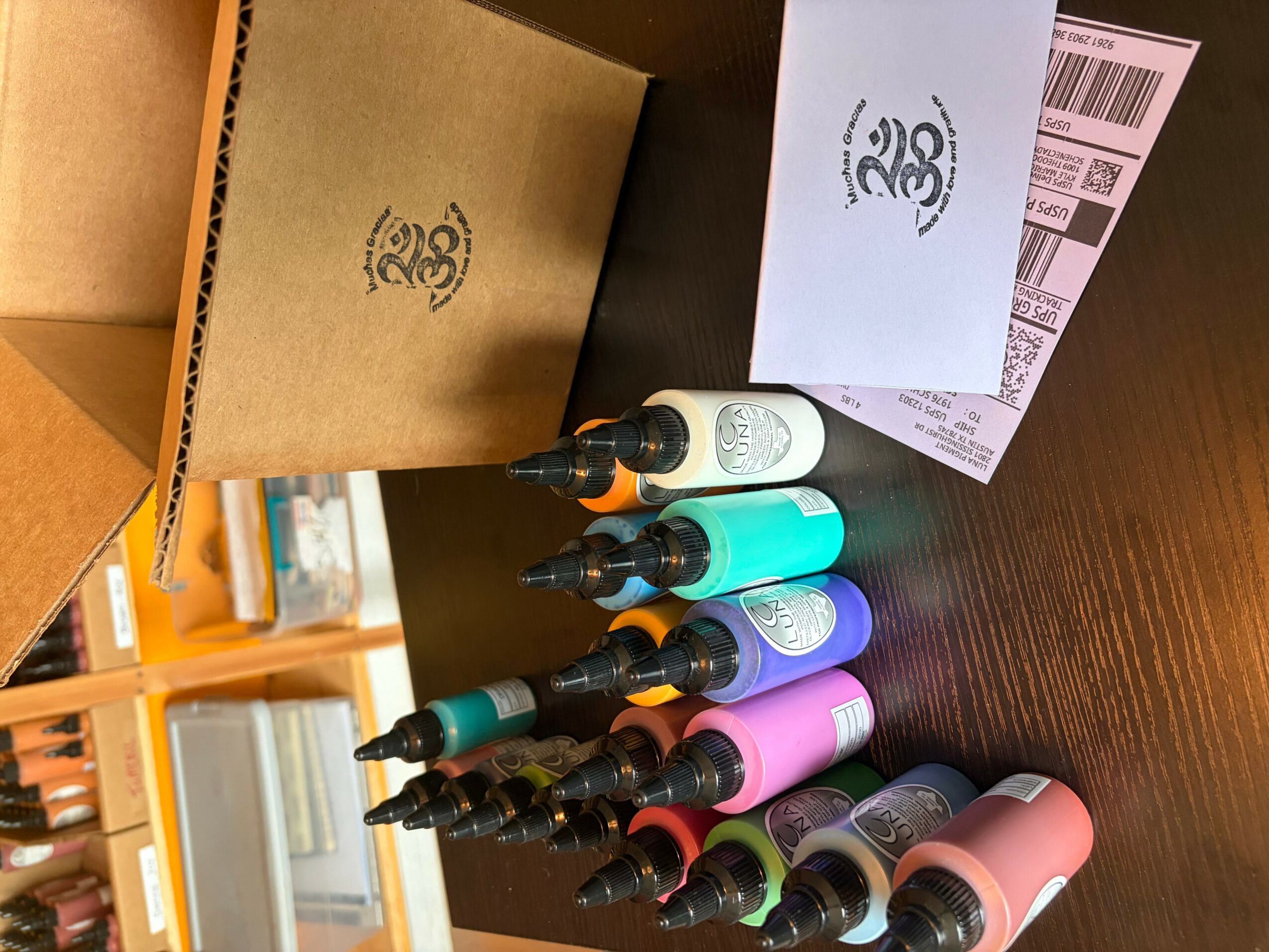 Six bottles of ink or paint arranged on a dark surface with packaging and paper nearby.