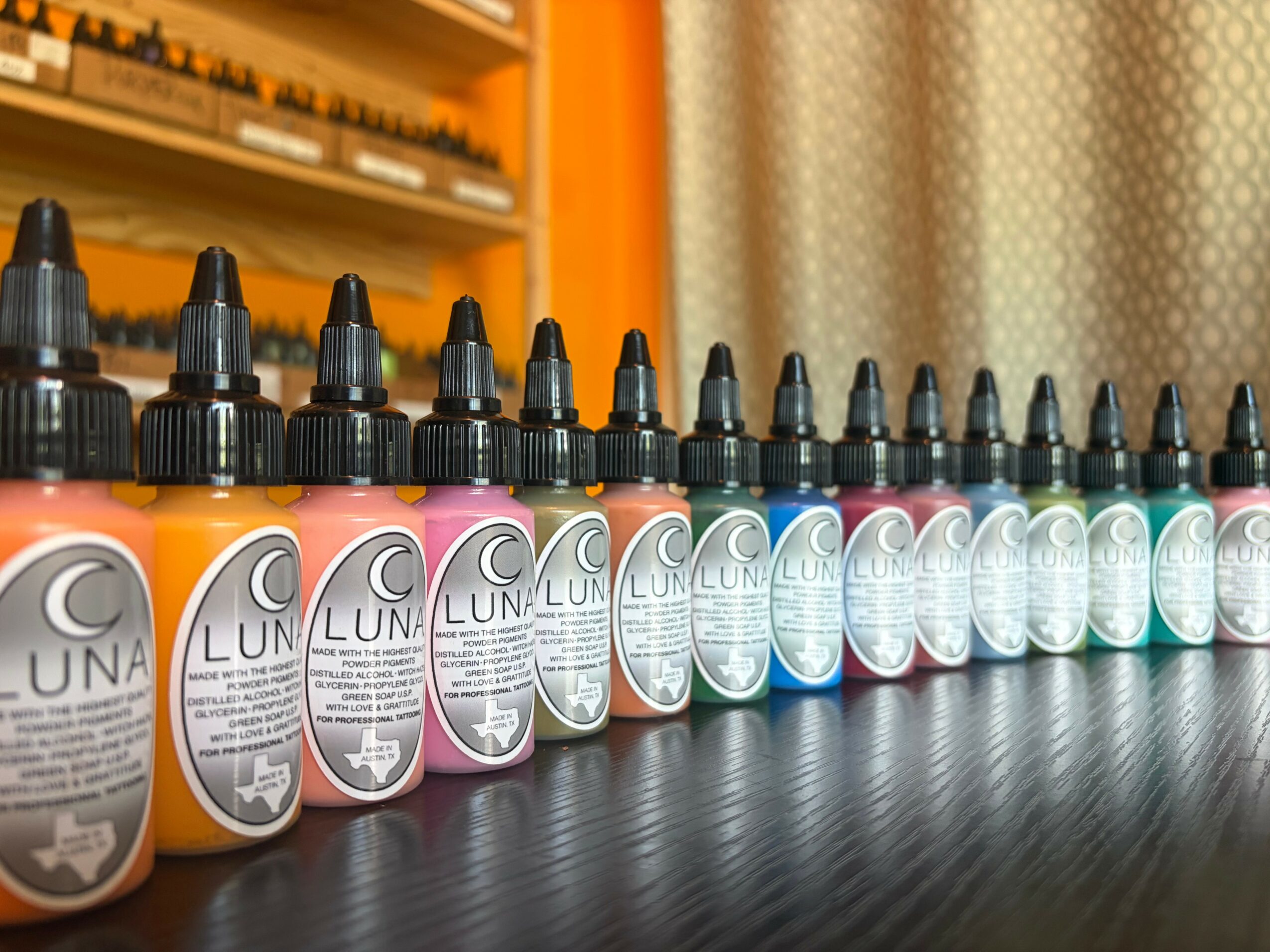 Row of colorful ink bottles with black caps on a dark surface, background of shelves and curtains.