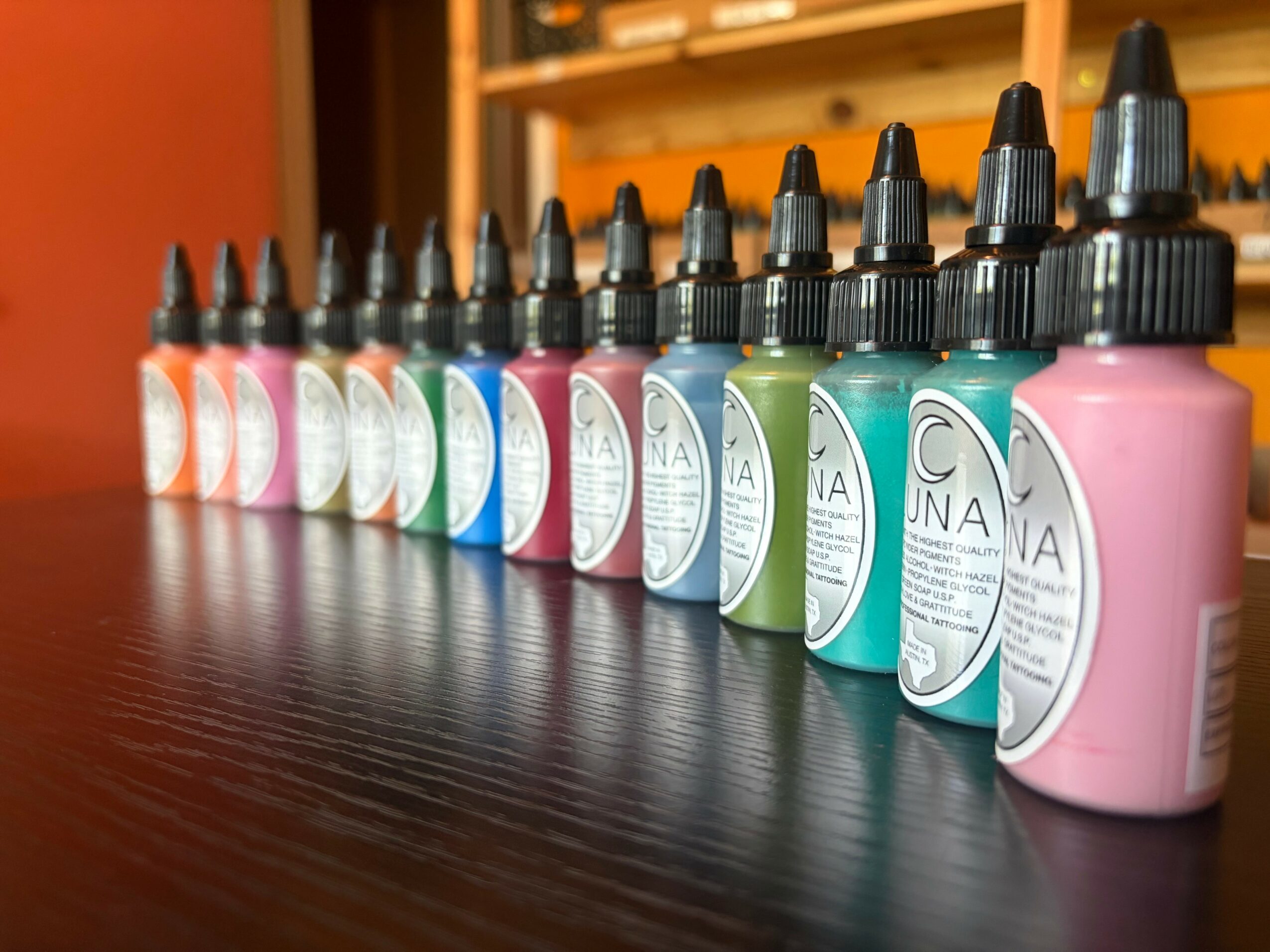 Row of colorful ink bottles with black caps on a dark surface, background of wooden shelves, vibrant colors, and labels visible.
