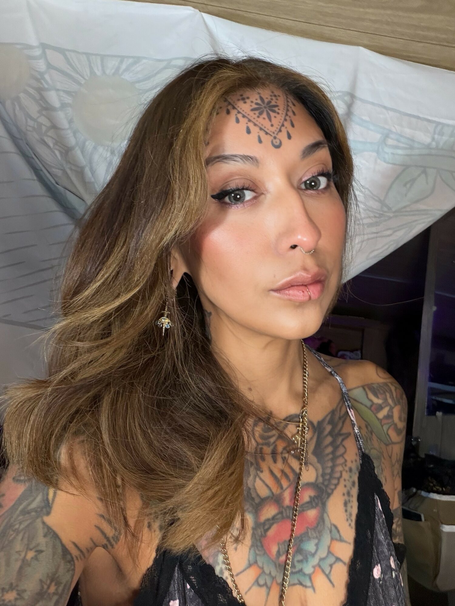 Woman with long wavy hair, makeup, and decorative face jewelry lying on a bed with patterned sheets, wearing a black top and jewelry.