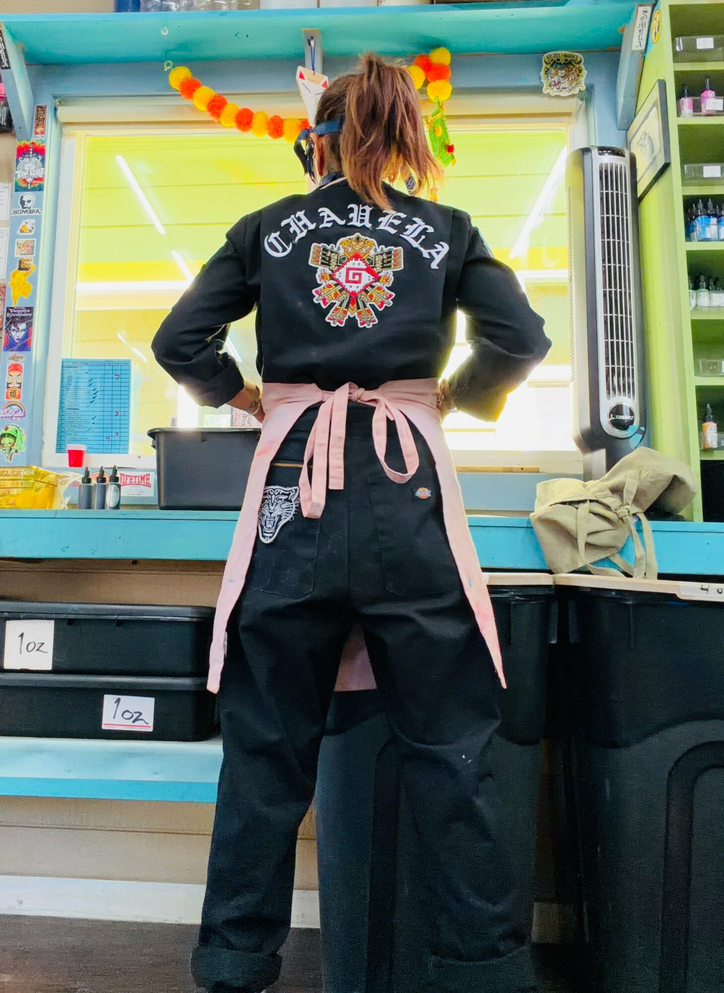 Person with hair in pigtails wearing a black jacket with a floral design, pink apron, standing in a colorful kitchen.