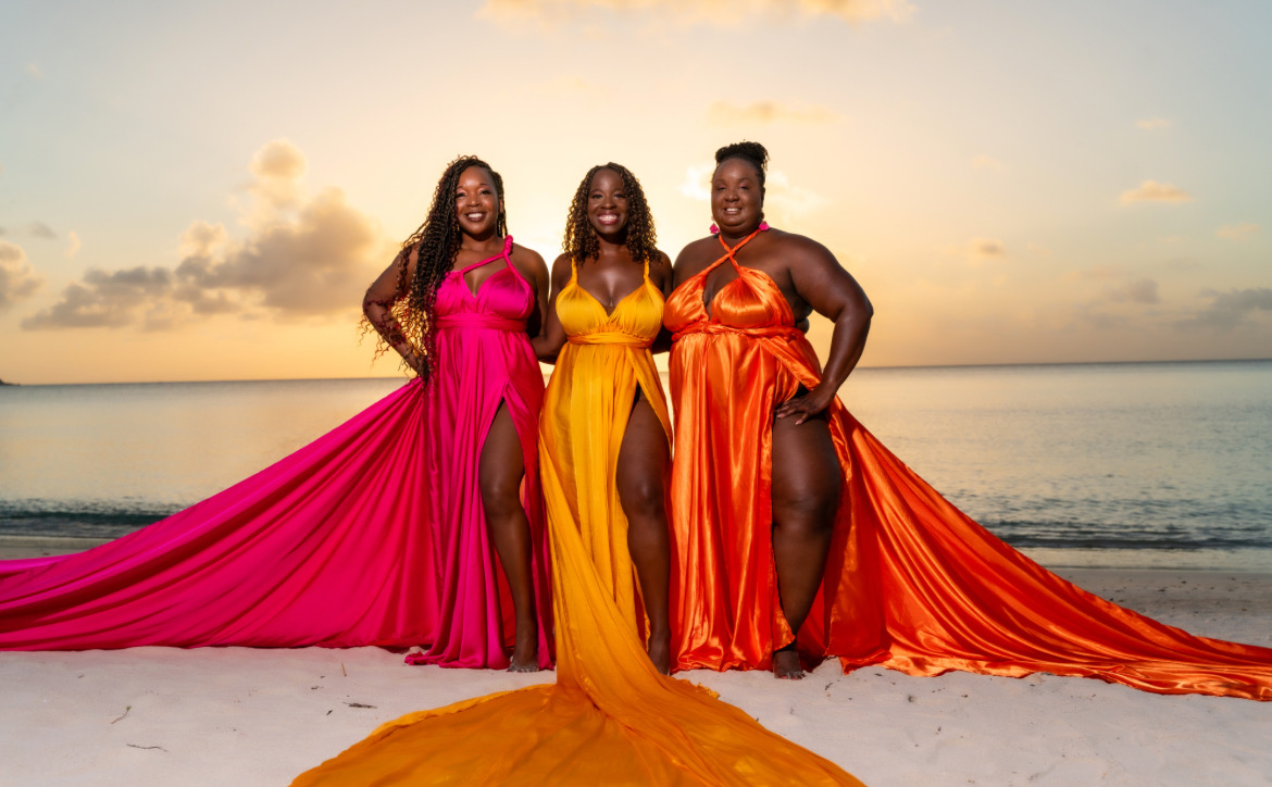 Three women in colorful dresses standing on a beach at sunset, with ocean and sky in the background.