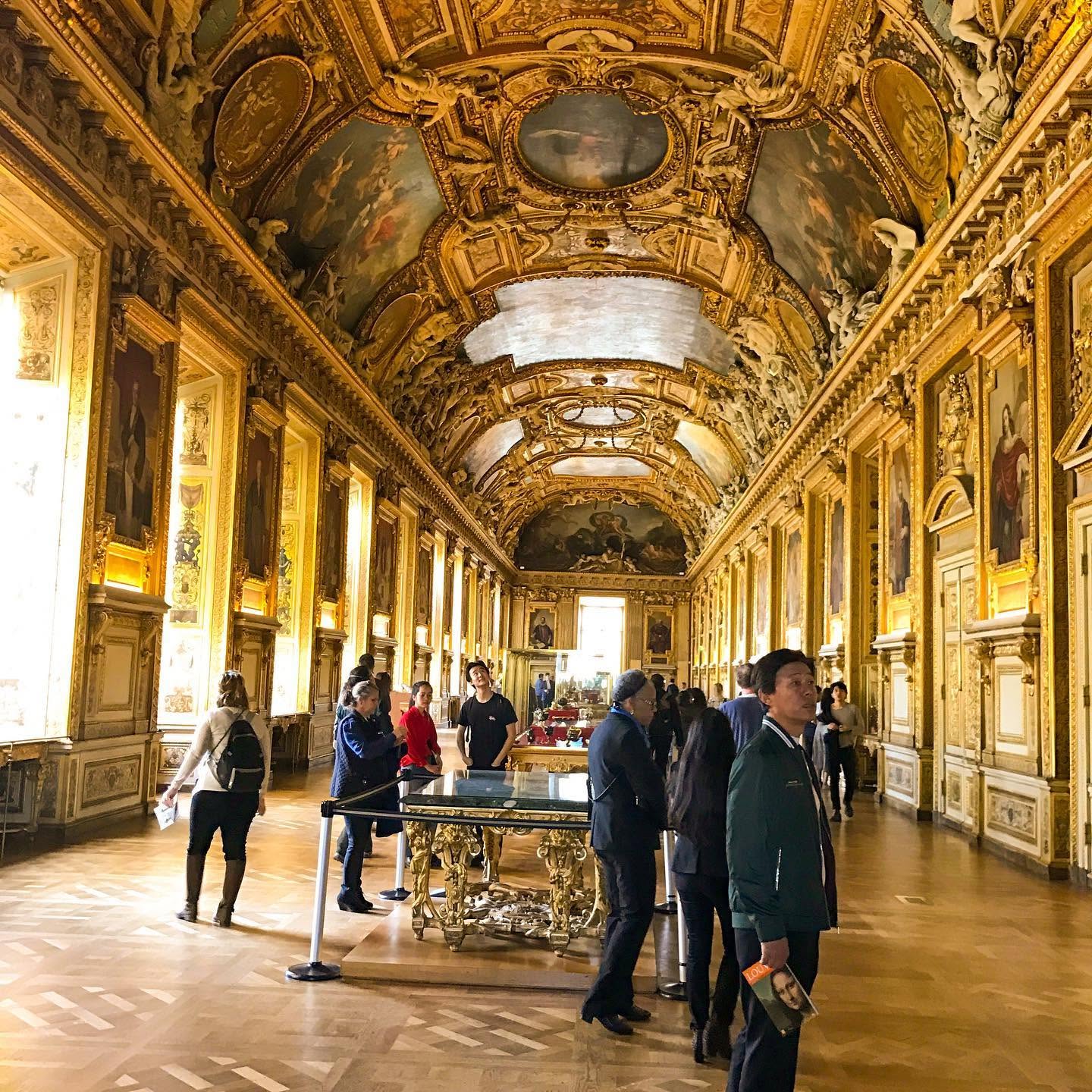 People inside a grand, ornately decorated hall with gold accents, paintings, and a high, decorated ceiling.