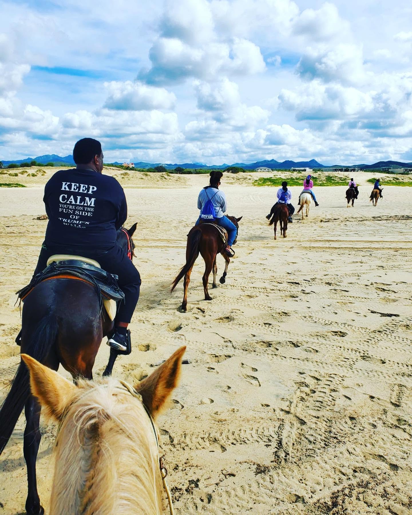 People riding horses on a sandy beach under a cloudy sky, with distant land and clouds in the background.